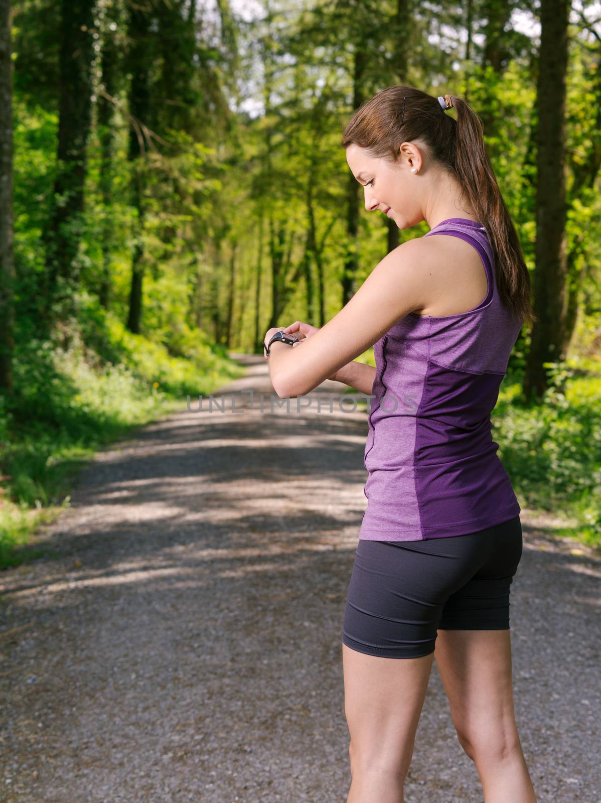 Photo of a young woman on a jogging path through a forest checking her running time.