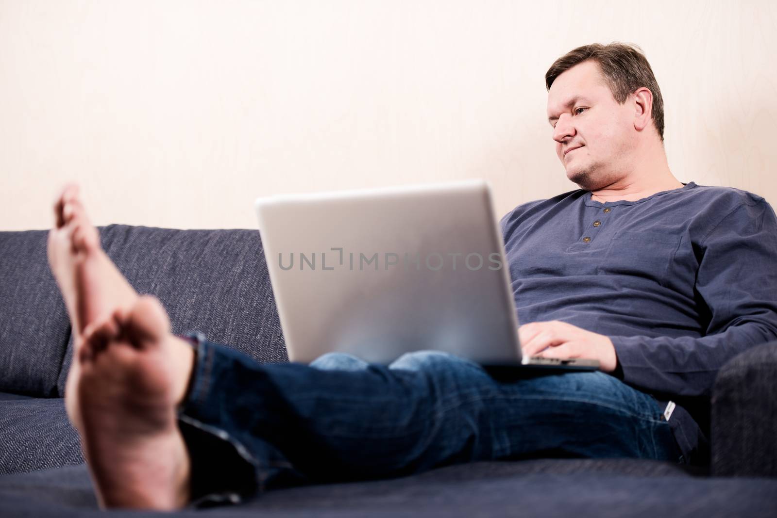 Man sitting on sofa with laptop barefoot by Nanisimova