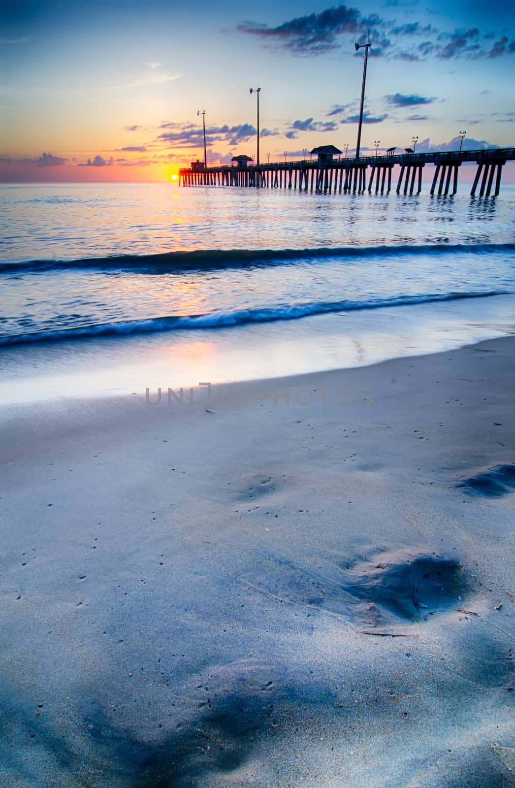 The rising sun peeks through clouds and is reflected in waves by the Nags Head fishing pier on the outer banks of North Carolina