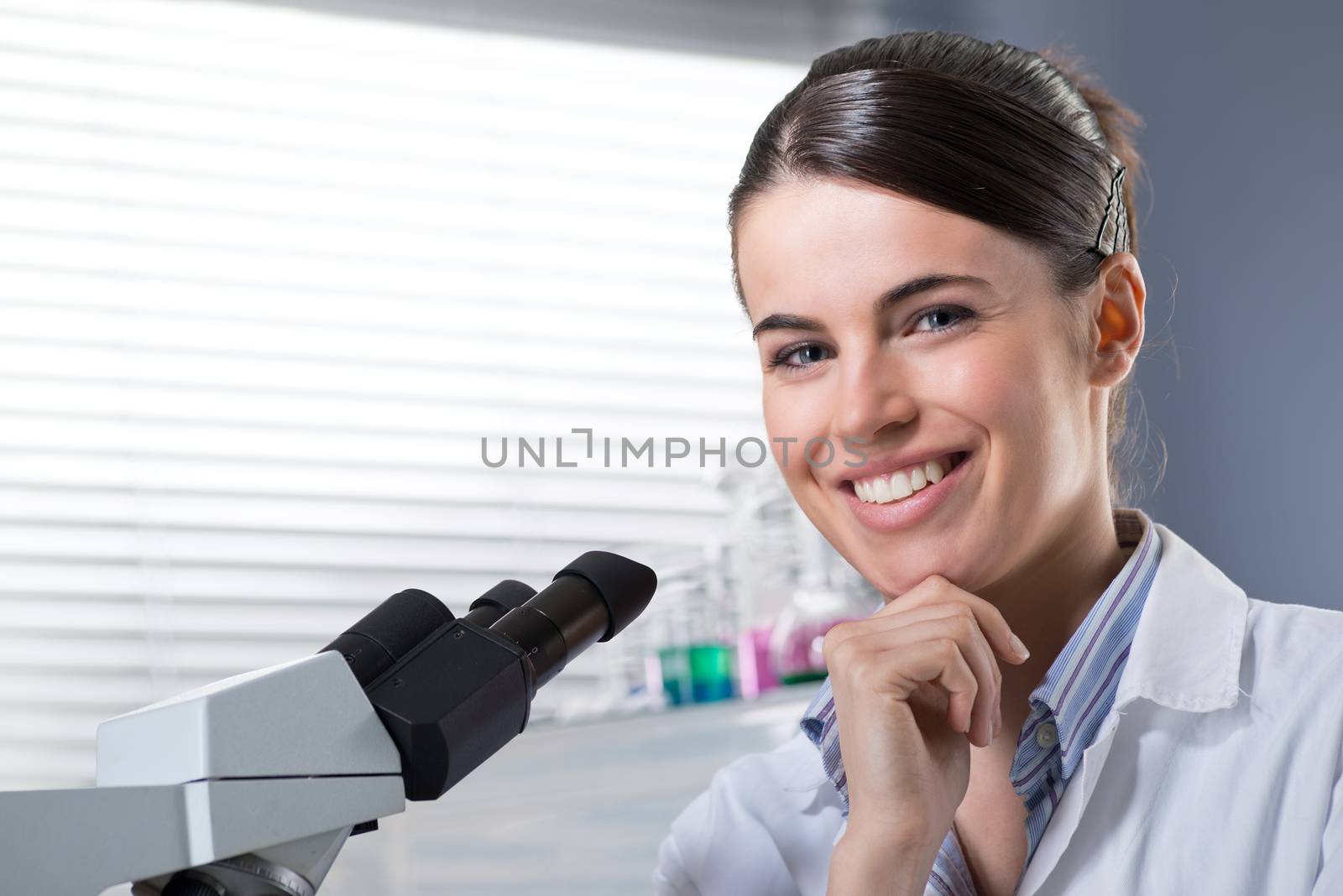 Confident female researcher in the chemistry lab with microscope and laboratory glassware on background.