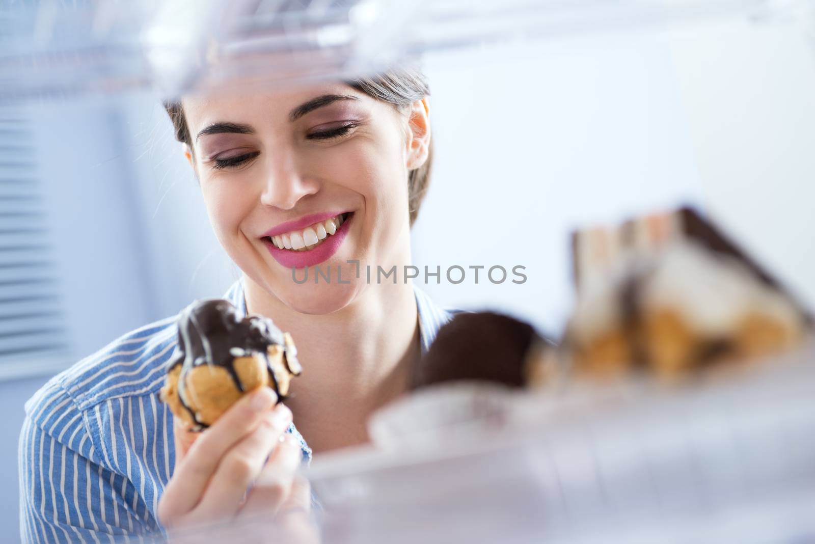Young woman taking pastry with chocolate topping from fridge and smiling. 