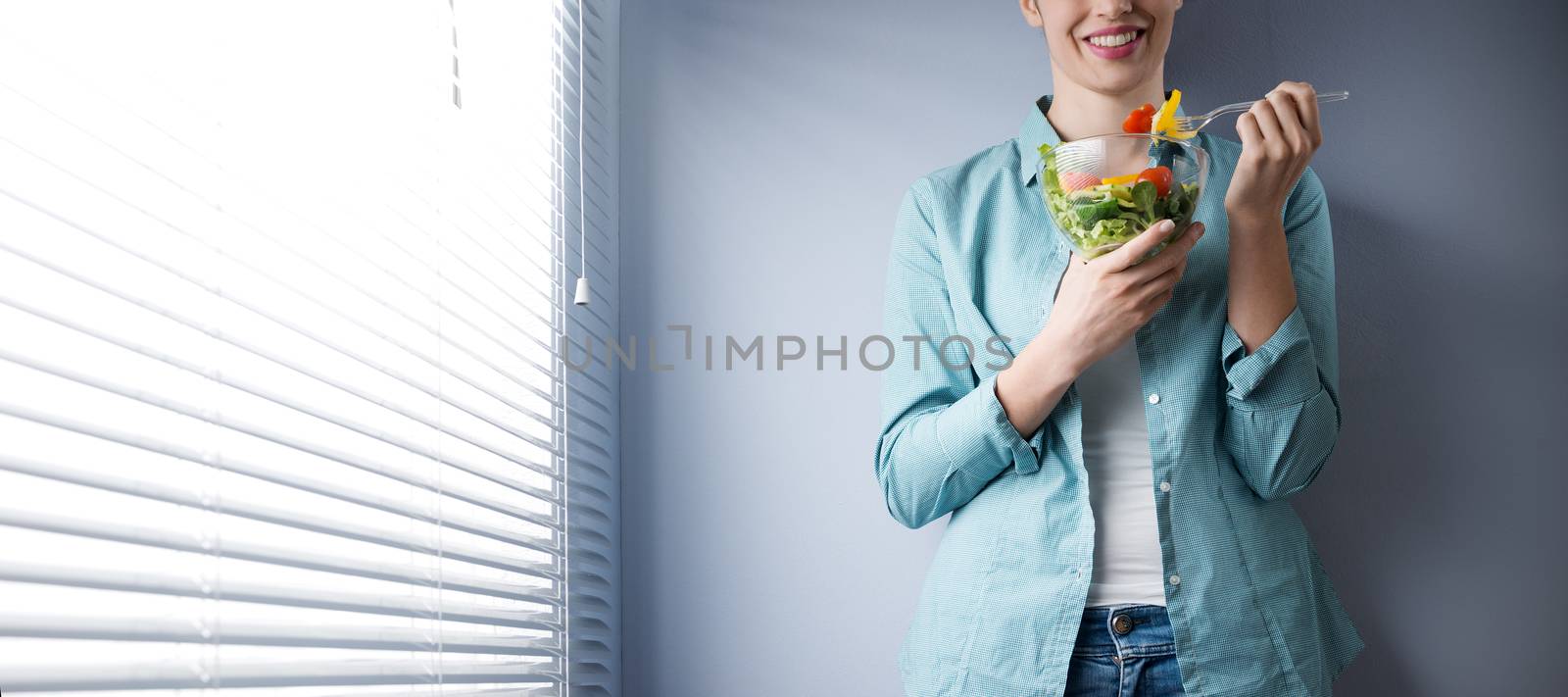 Woman smiling and eating salad in front of a window.