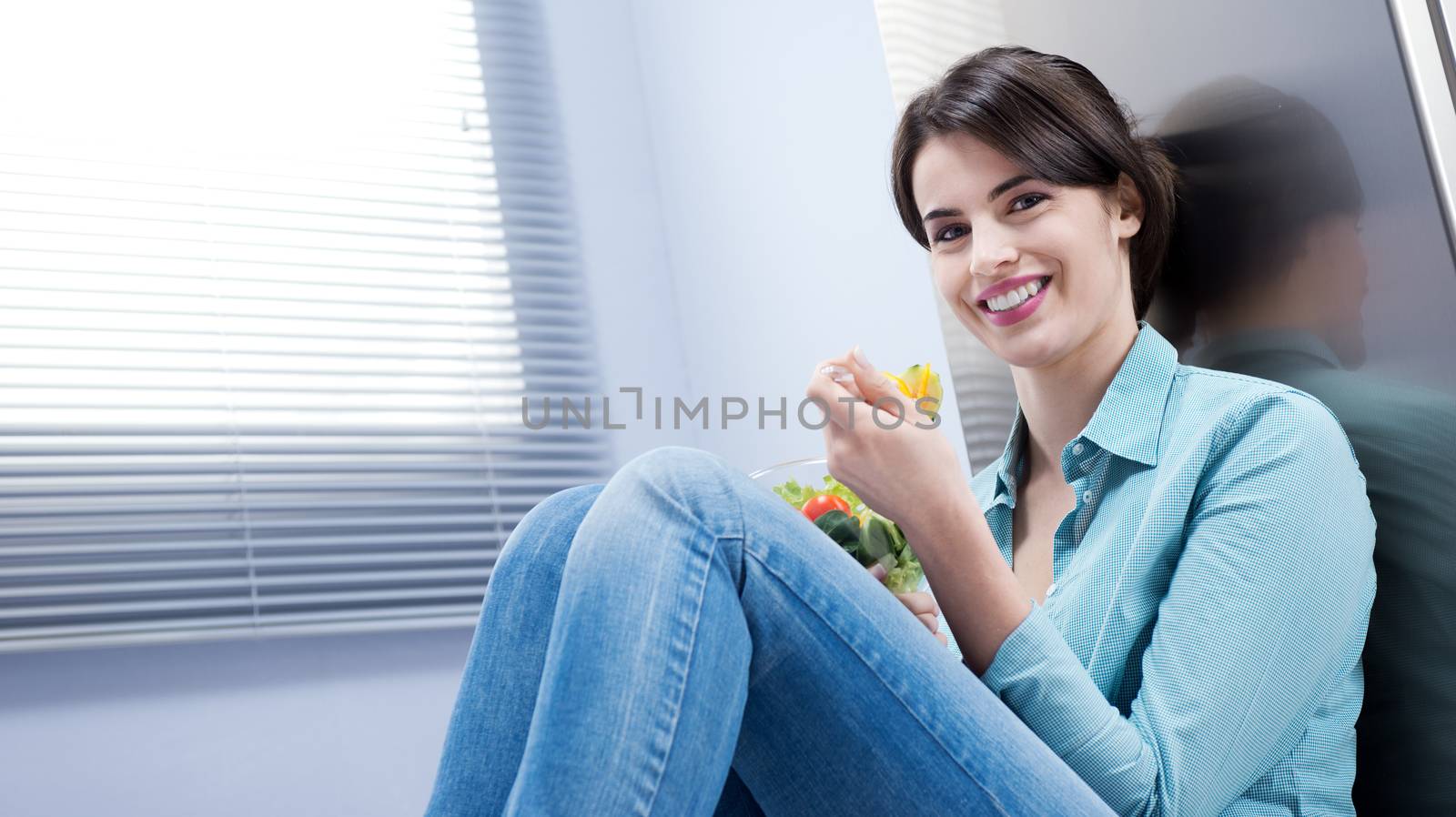 Young woman sitting in front of the fridge eating salad.