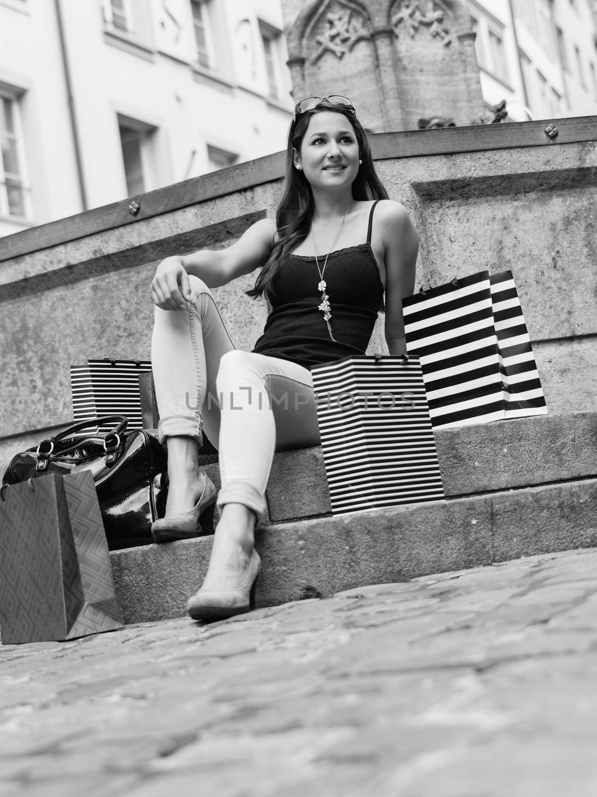 Photo of a beautiful young woman sitting and resting with her shopping bags at a fountain in an old European city.