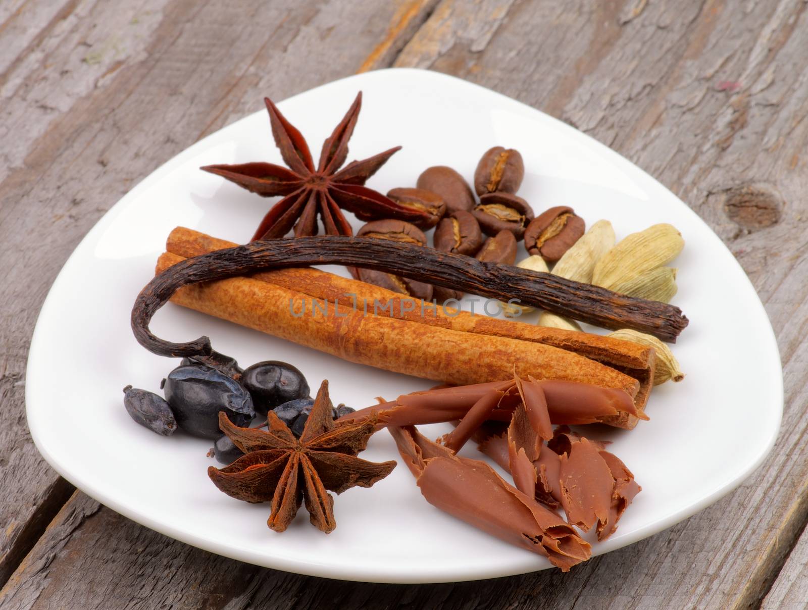 Arrangement of Chocolate Slices, Vanilla Pods, Anise Stars, Cardamon and Coffee Beans on White Plate isolated on Wooden background