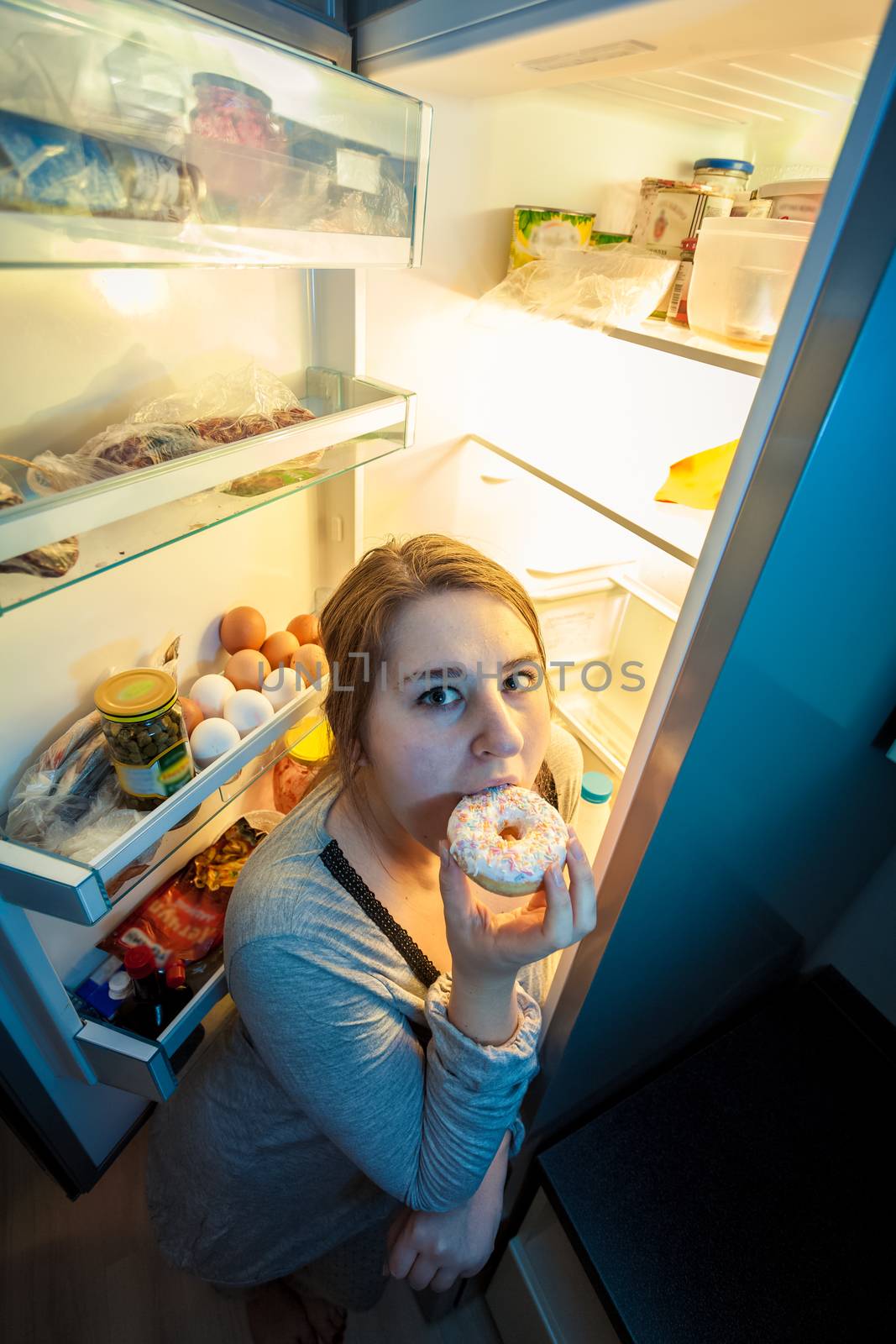 Portrait of young woman in pajamas eating donut next to refrigerator