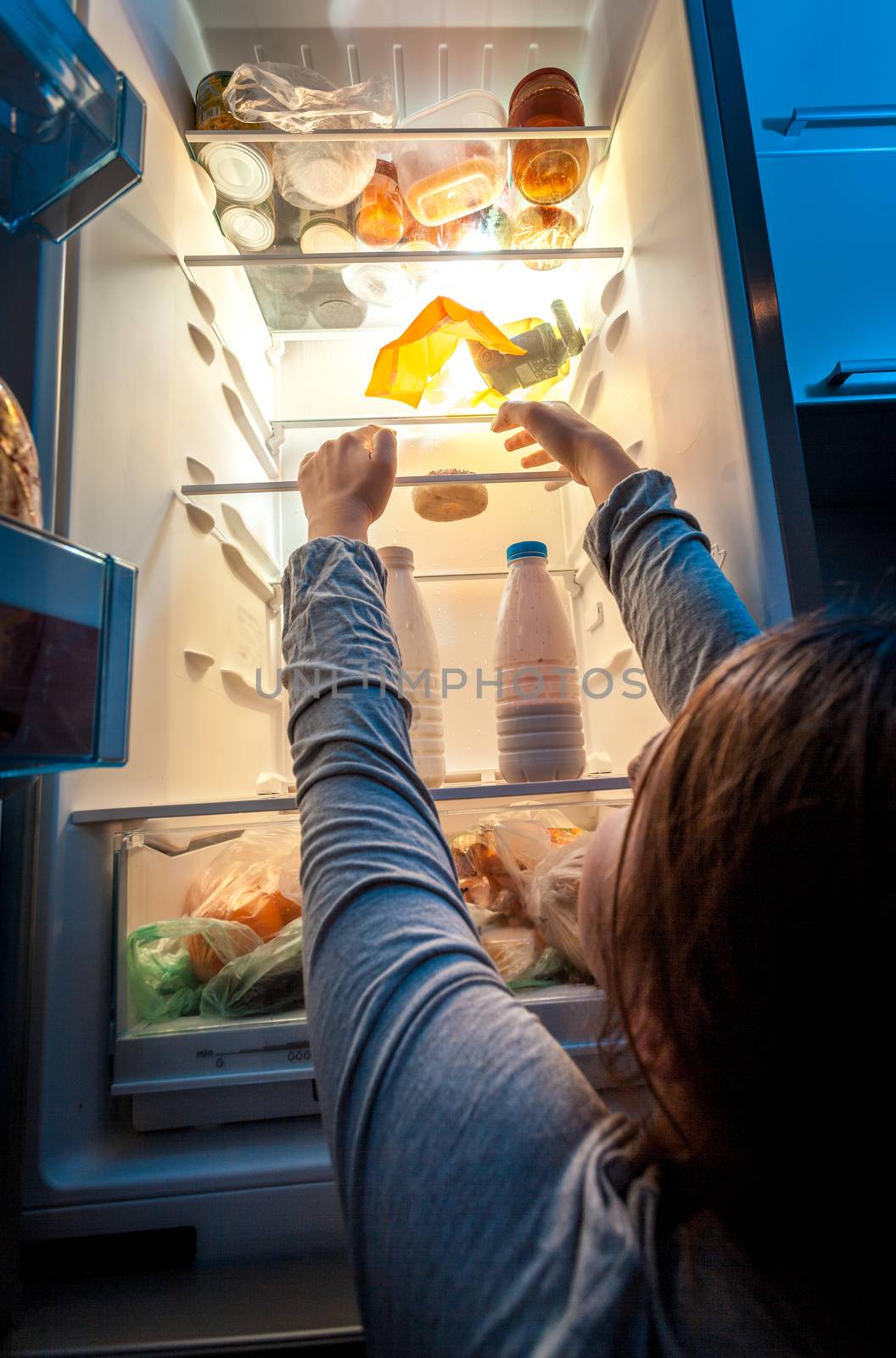 Portrait of woman in pajamas reaching donut on top shelf of fridge at night