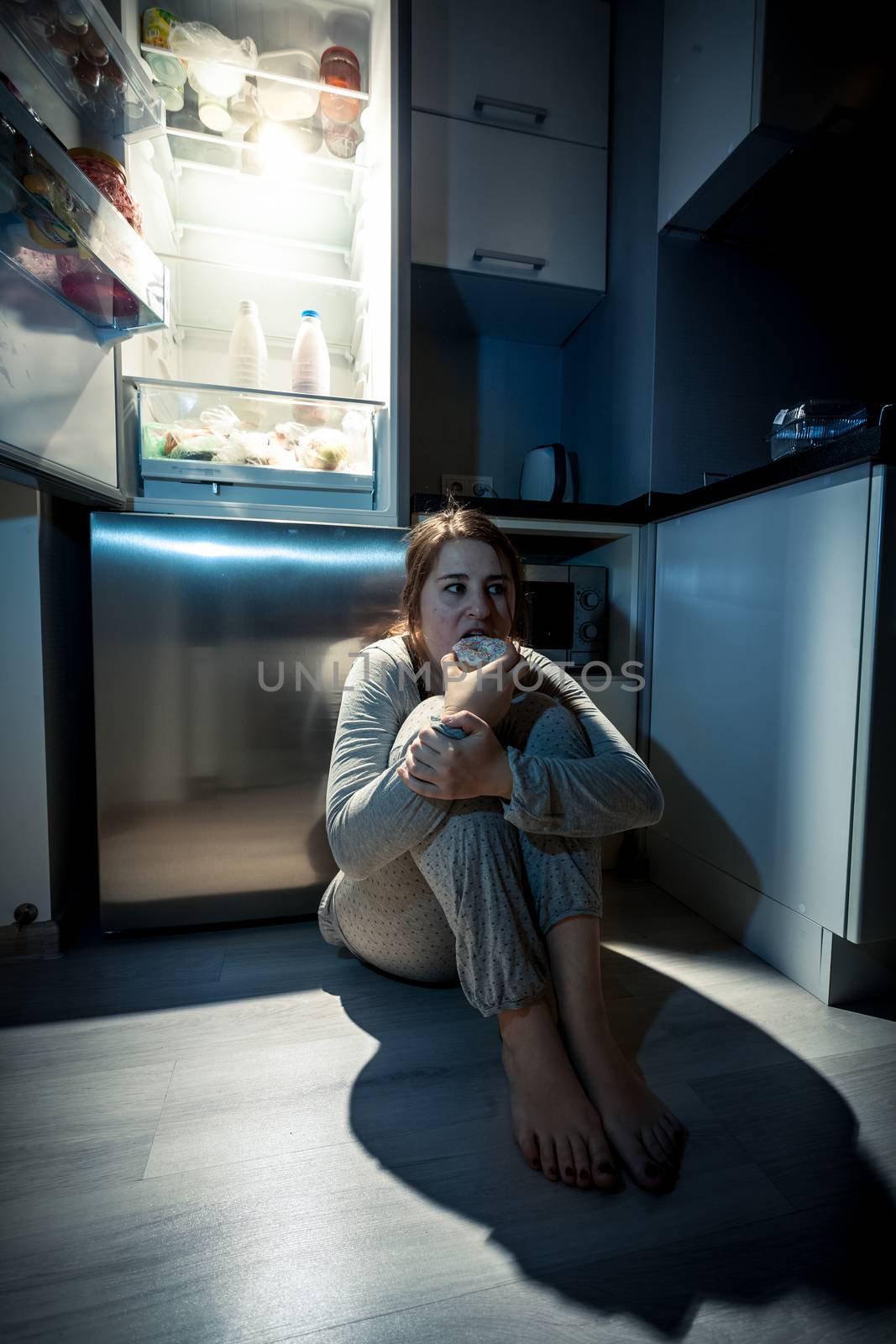 Photo of young woman eating next to refrigerator at night