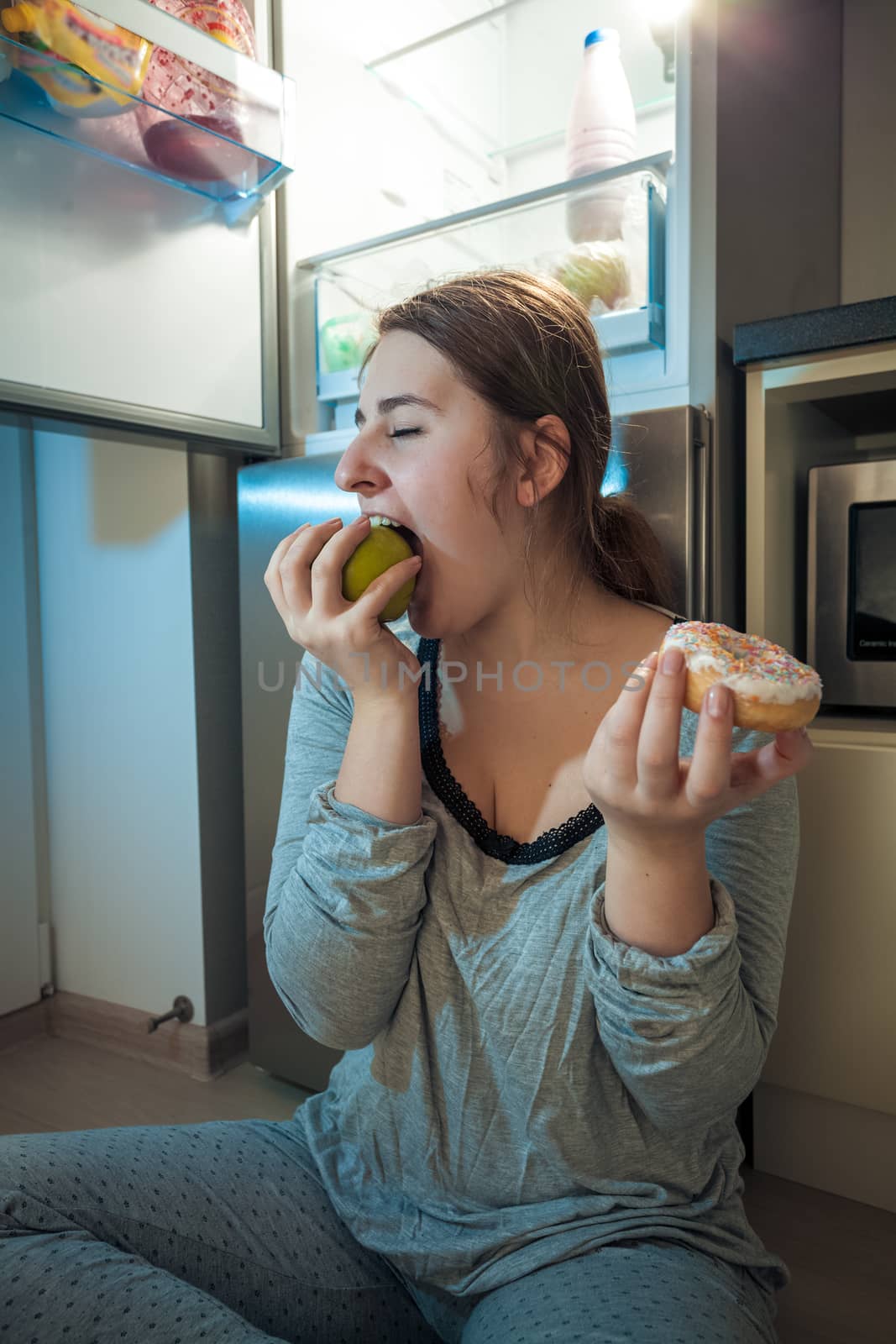 Closeup portrait of woman in pajamas biting apple at kitchen at night