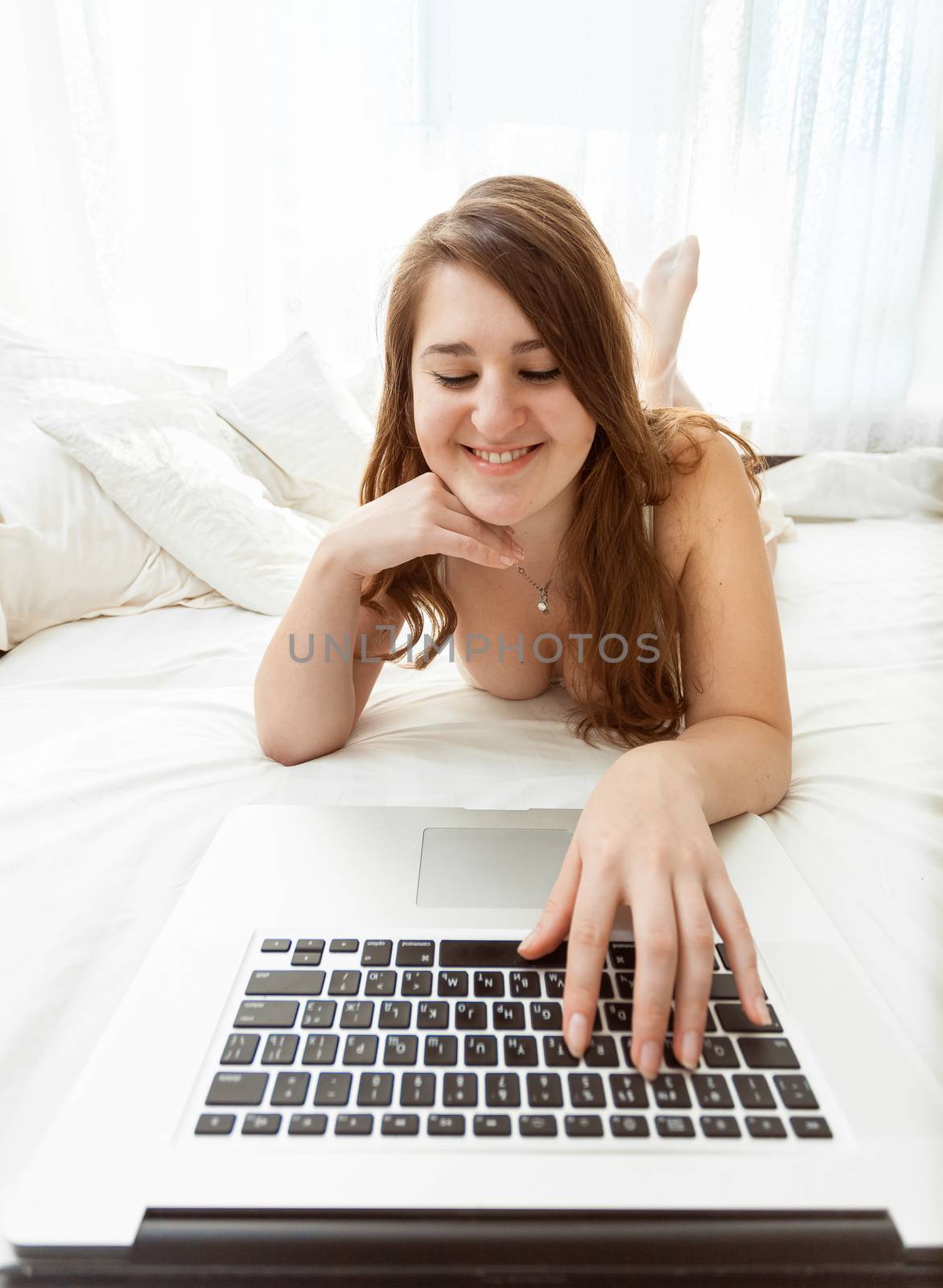 Wide angle portrait of sexy woman lying on bed and typing on keyboard