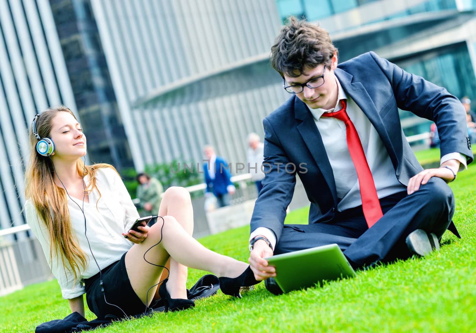 Successful Junior executives dynamics laying down on green grass near a modern glass office building, listening music and using laptop
