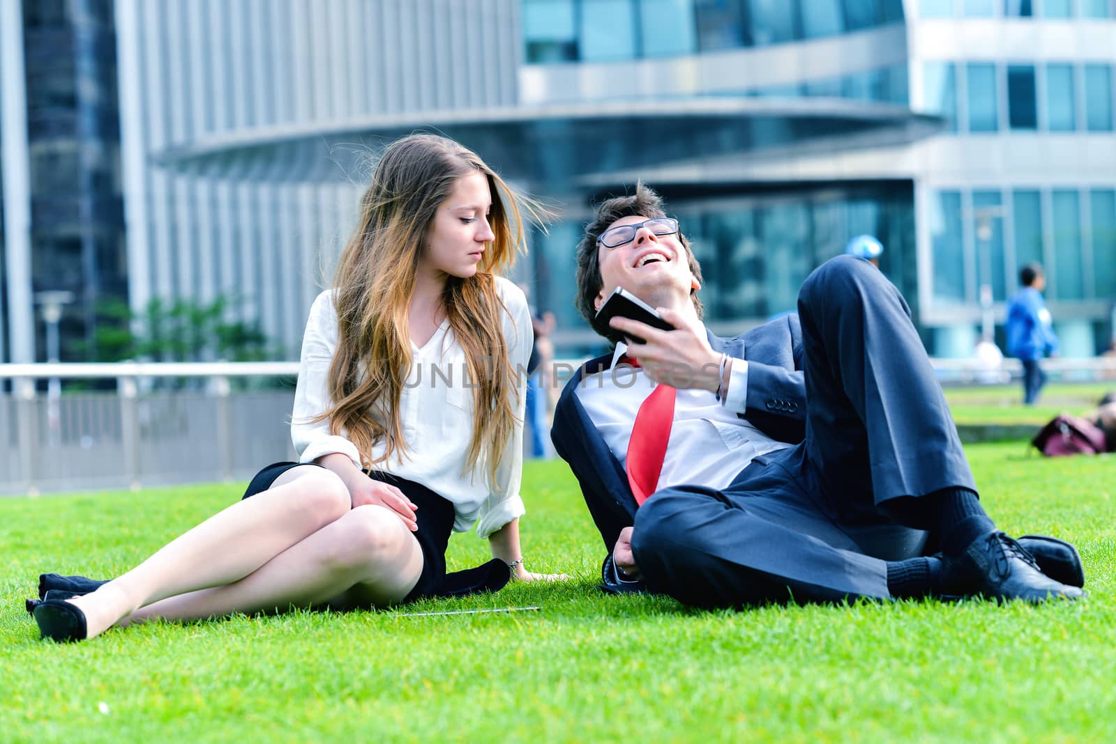 Successful Junior executives dynamics laying down on green grass near a modern glass office building, listening music on smartphone