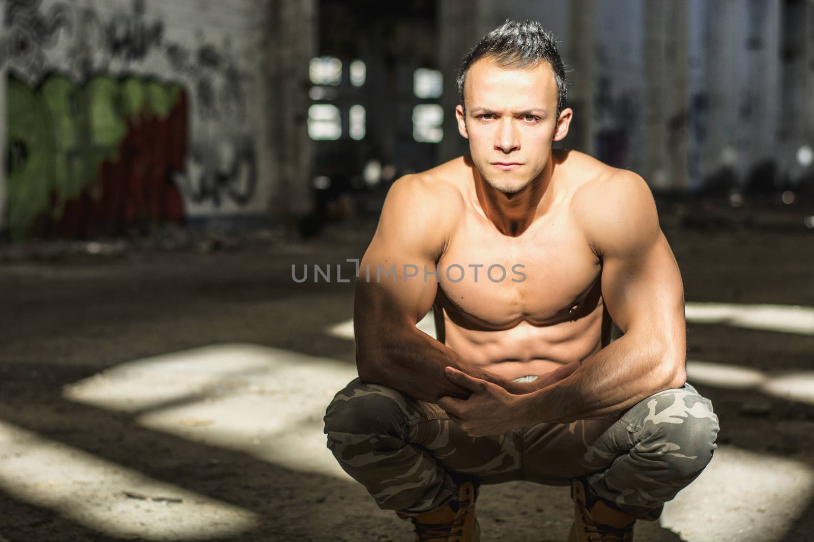 Muscular shirtless young man in abandoned warehouse sitting, looking at camera