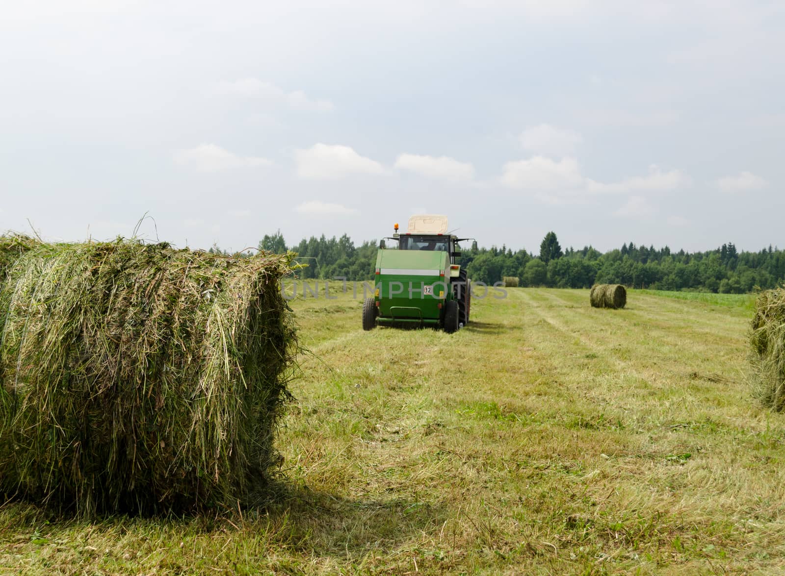 Straw bales and agricultural machine tractor collect gather hay in field near rural village houses.