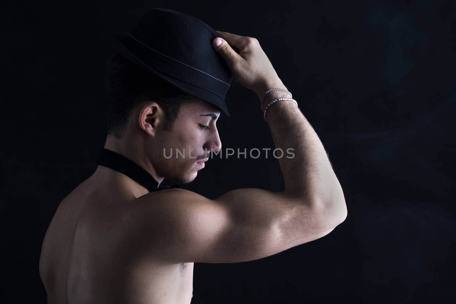 Profile of shirtless young latino man holding black fedora hat, on dark background