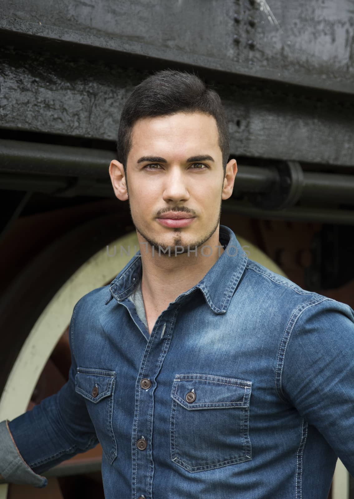 Handsome young man in denim shirt in front of old train, looking at camera