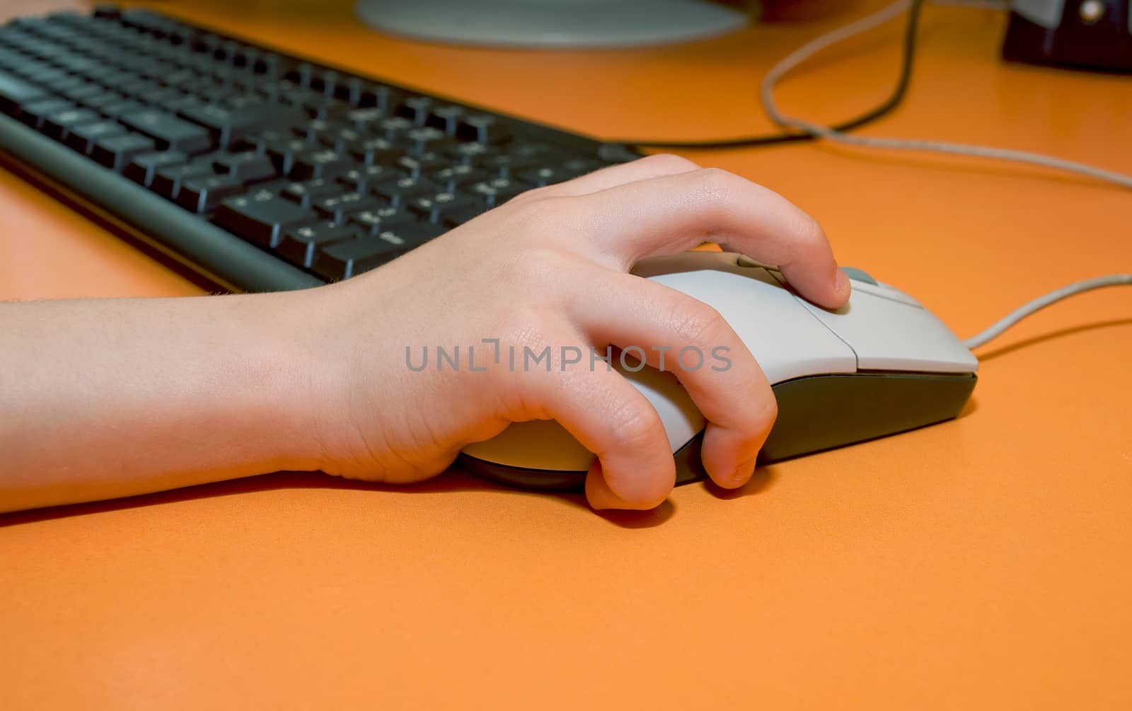 Children's right hand on the mouse on the background of computer keyboard on the orange table