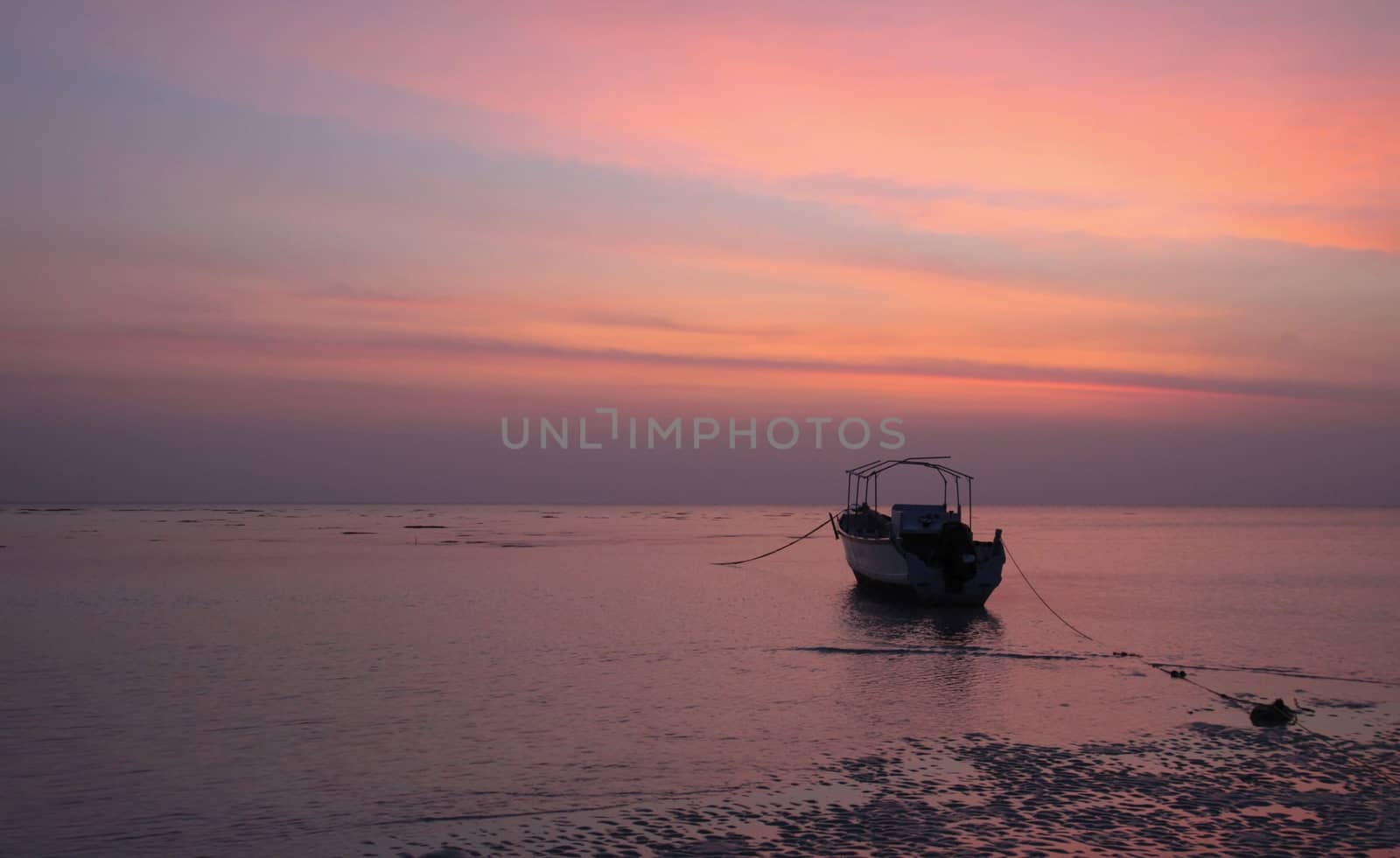 The low tide of predawn and dead calm sea immobilises this lone boat anchored near shoreline while the approaching dawn is reflected by the shades of blue and orange in the sky.