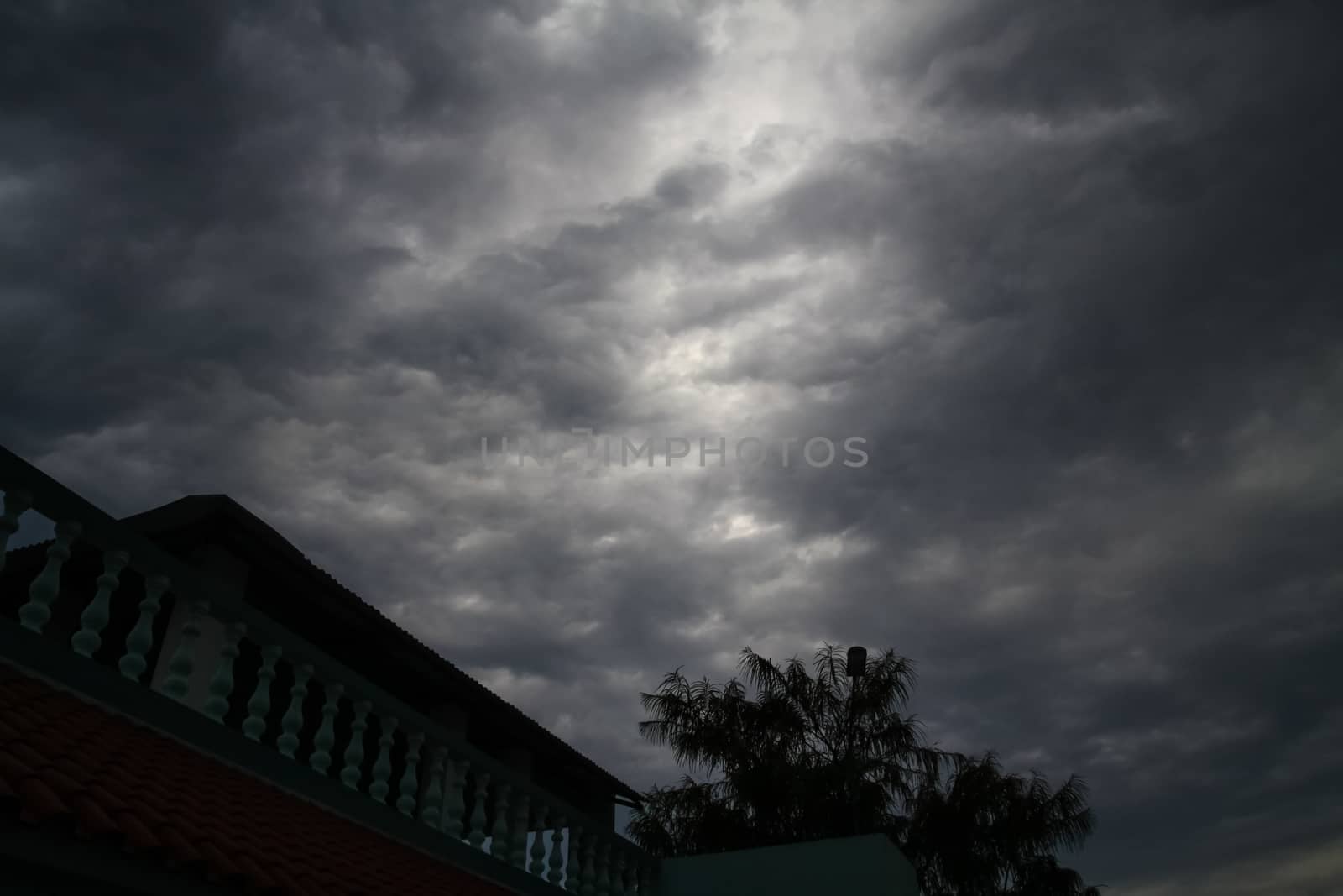 A view of dark menacing storm clouds encompassing the gray scale spectrum with part of the housetop and a tree top in the foreground providing a sense of scale