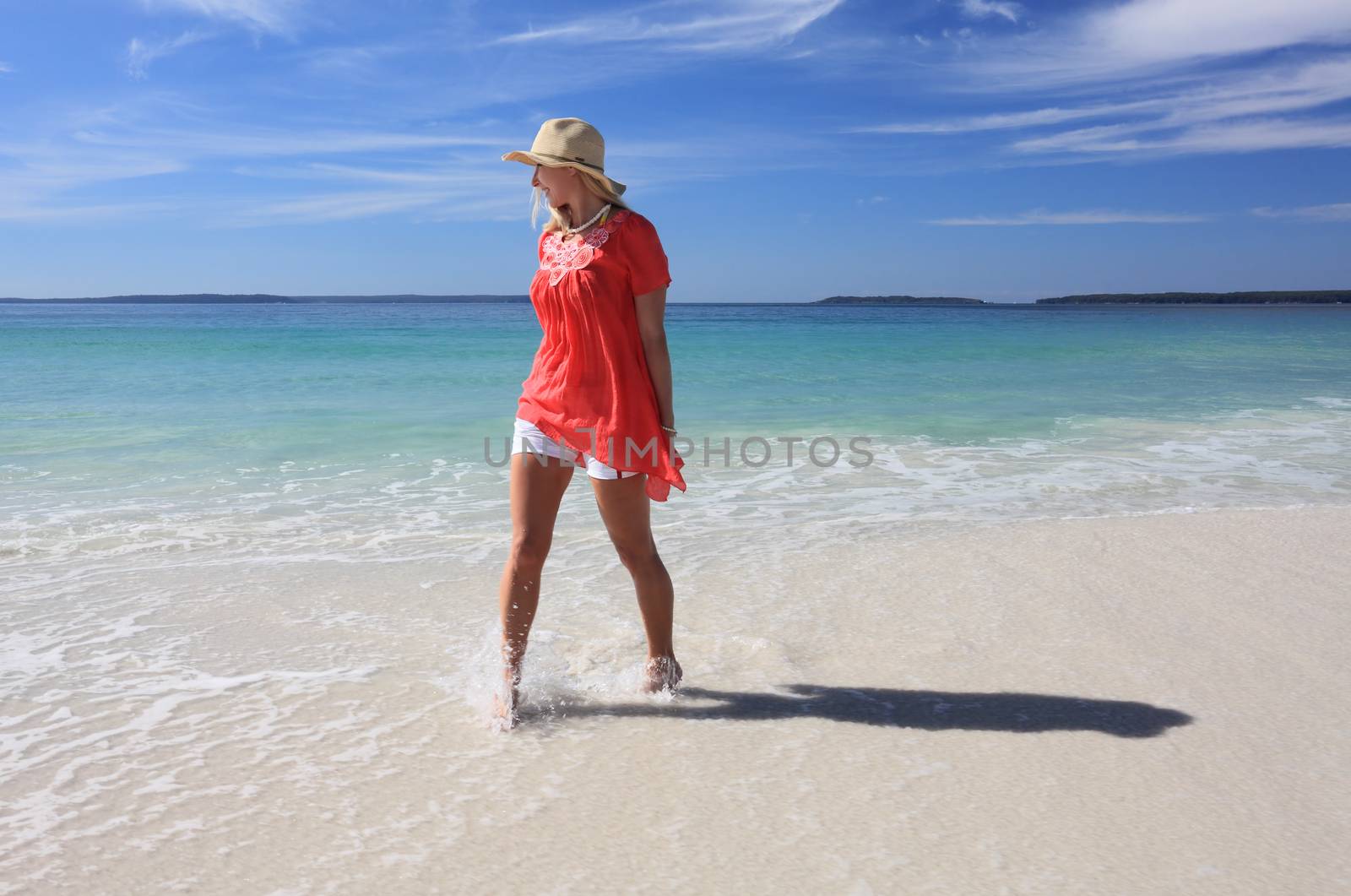 Happy smiling woman wearing a sun hat splashing feet while walking along the beach.   Some motion in splashing water and feet.