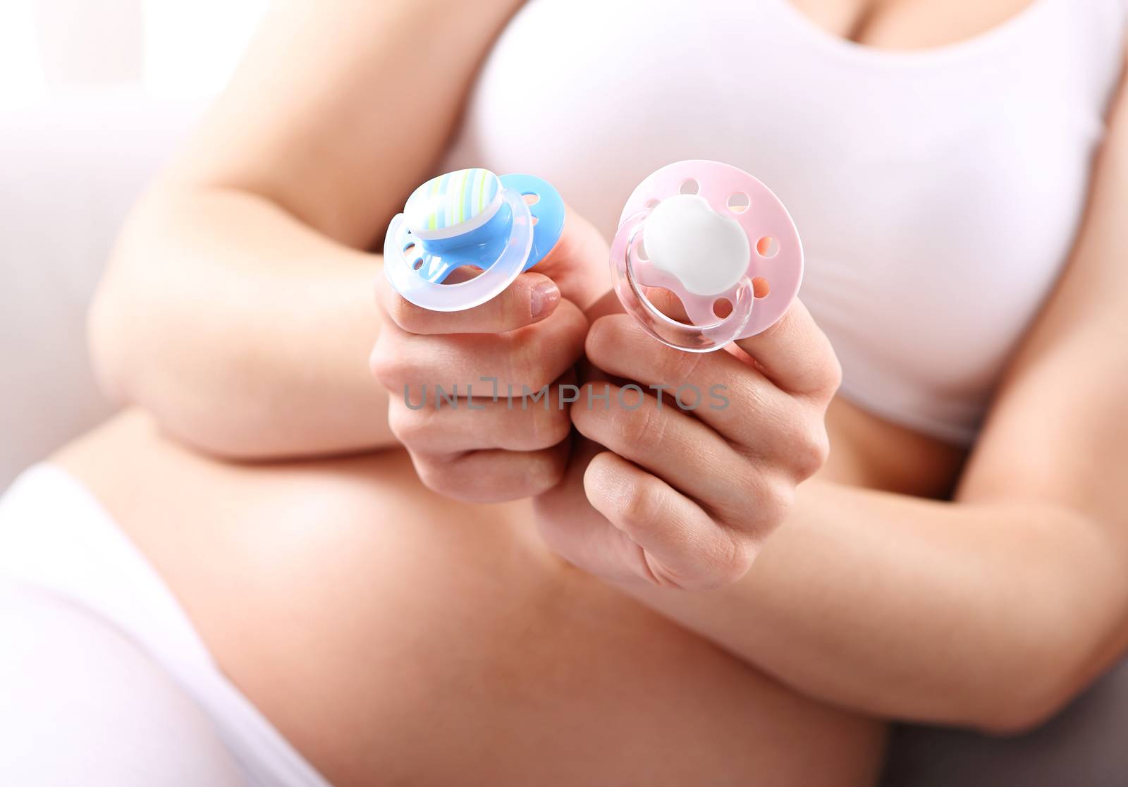 A pregnant woman is holding in his hand the two children's pacifiers blue and pink.