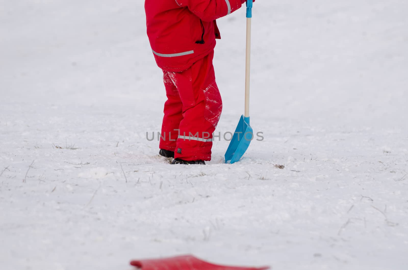 child stand with large snow shovel dressed in red waterproof clothing outdoor in winter time