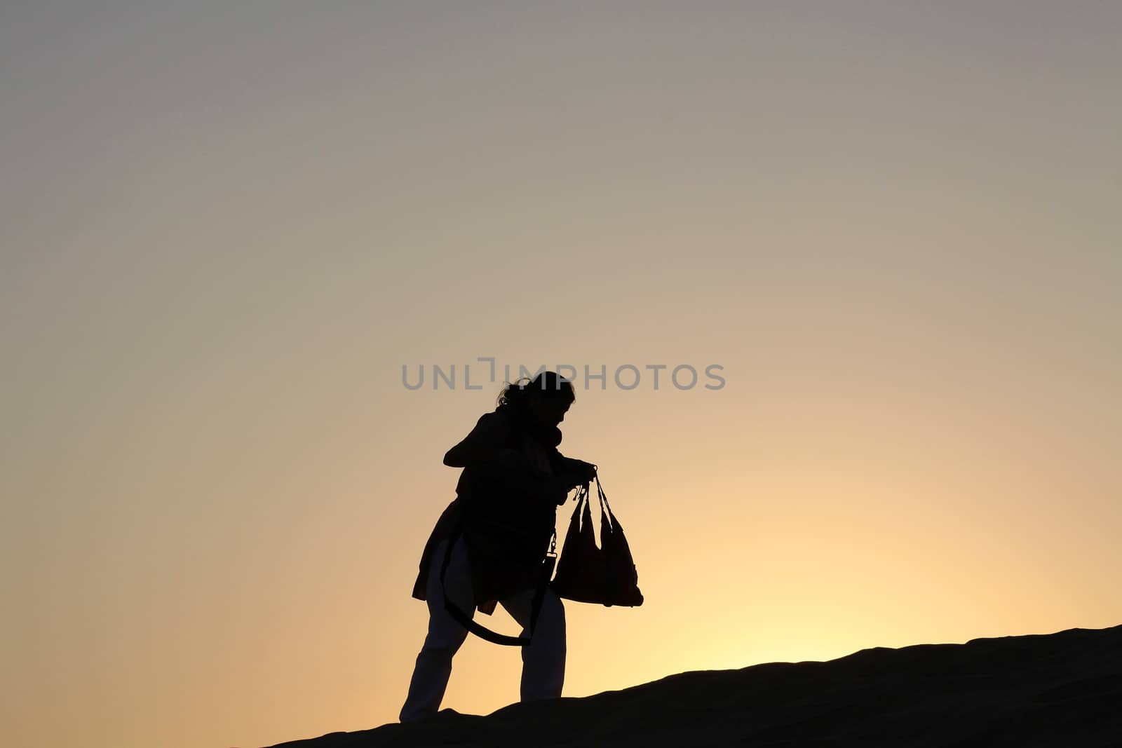 Single Woman walking uphill is silhoutted by the bright orange hues of the Dusk Twilight. Her posture perfectly conveys the peoples eternal struggles to overcome natures obstacles