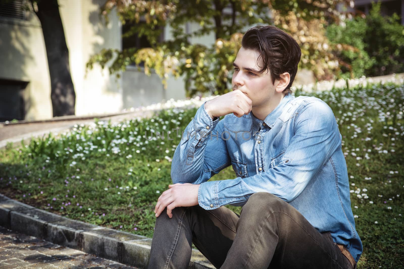 Young man sitting outdoors in public park, with hand on his chin looking away thinking