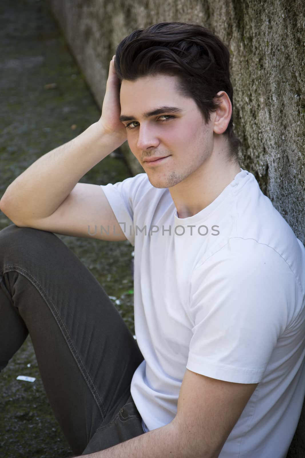 Attractive young man sitting on the ground against rock outdoors, looking at camera with a smile