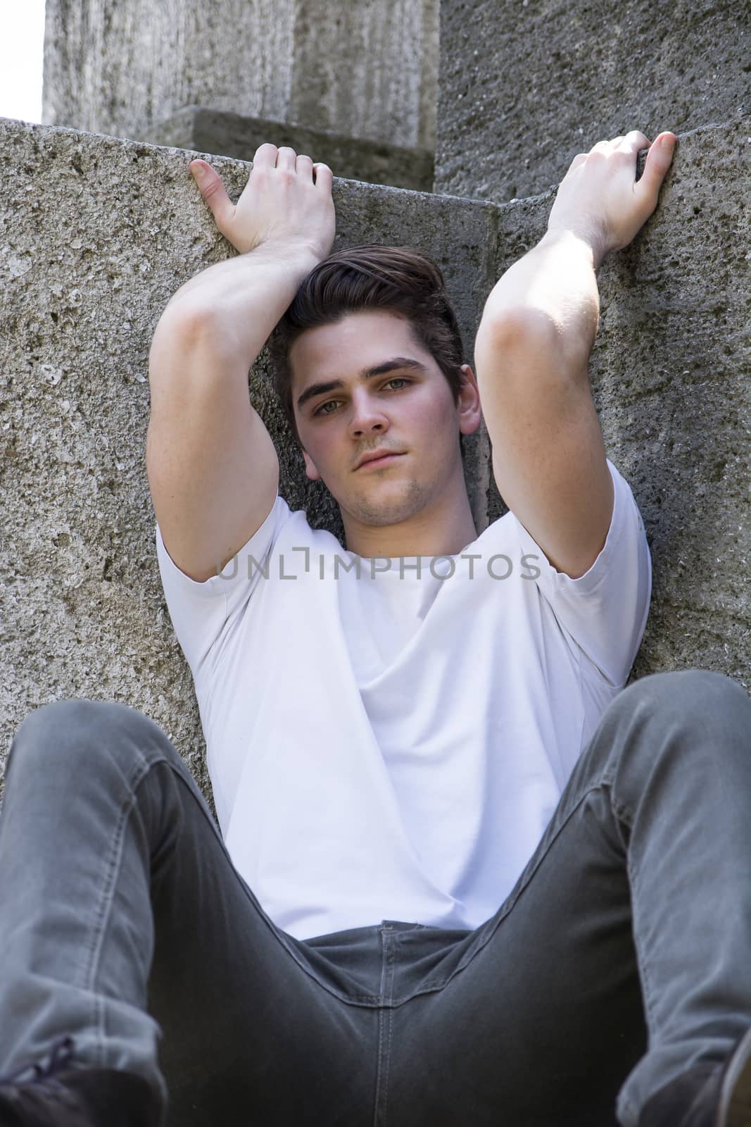 Attractive young man sitting on the ground against stone blocks outdoors, looking at camera, arms up