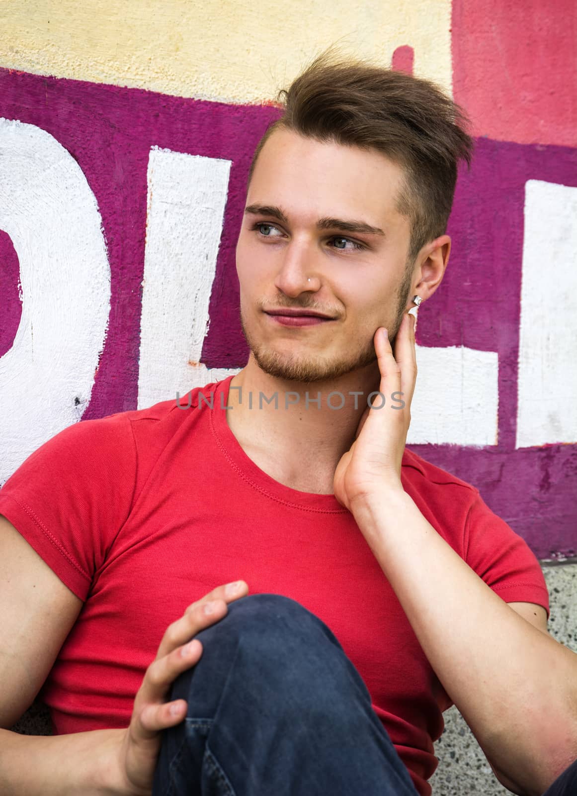 Handsome blond young man sitting against colorful graffiti wall looking away with a smile