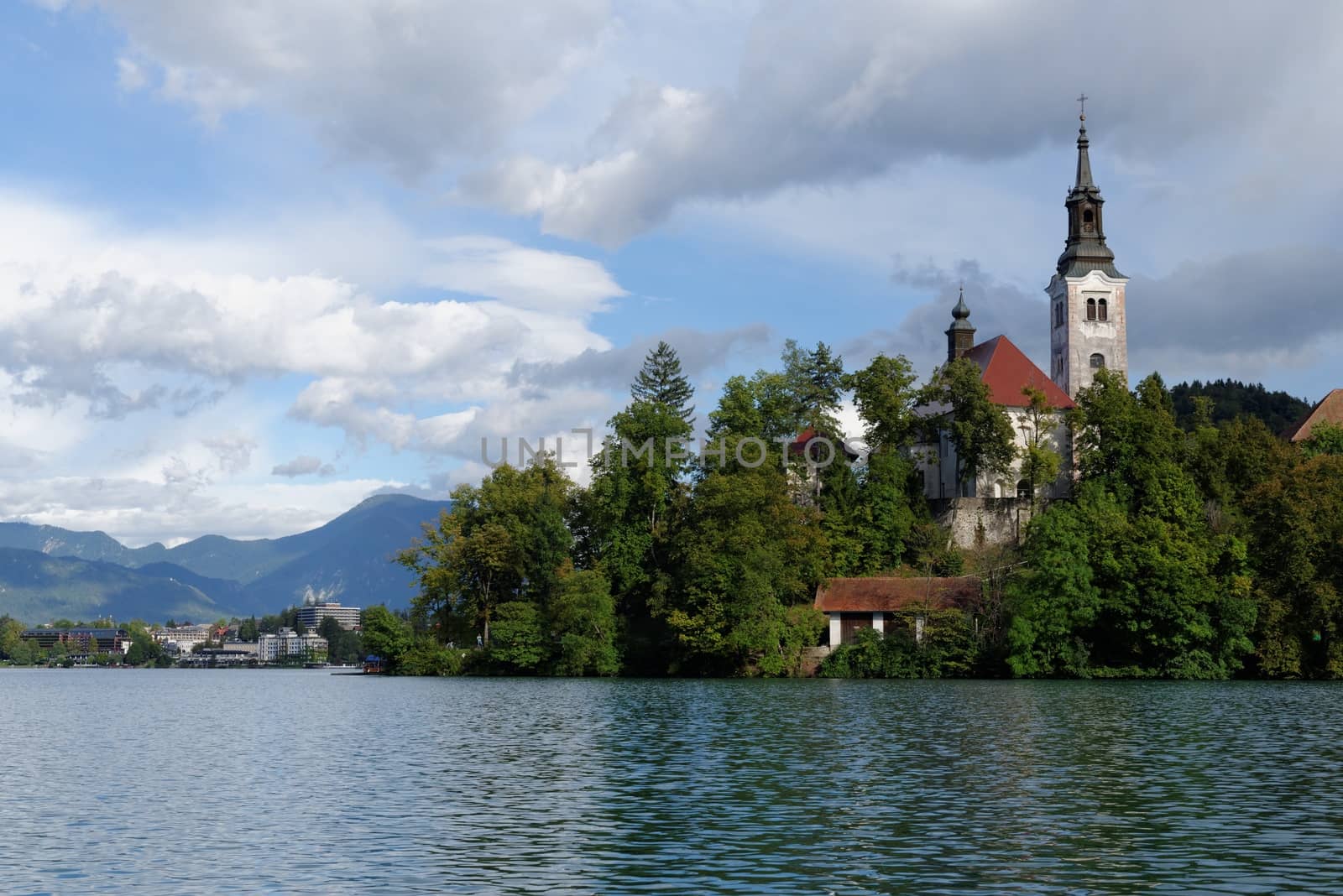 Catholic church situated on an island on Bled lake with mountains and resort on the background 