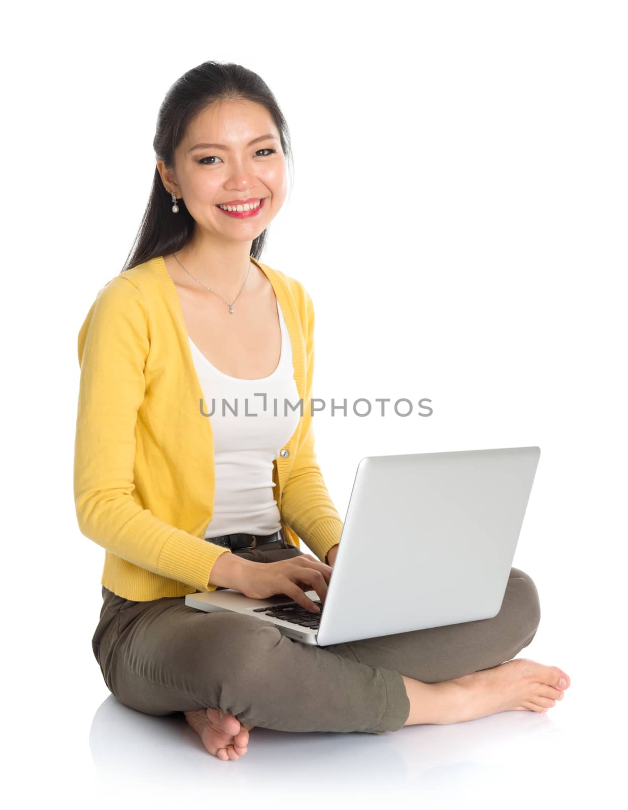 Full body Asian woman in yellow blouse using laptop computer notebook, looking at camera, seated on floor isolated on white background.