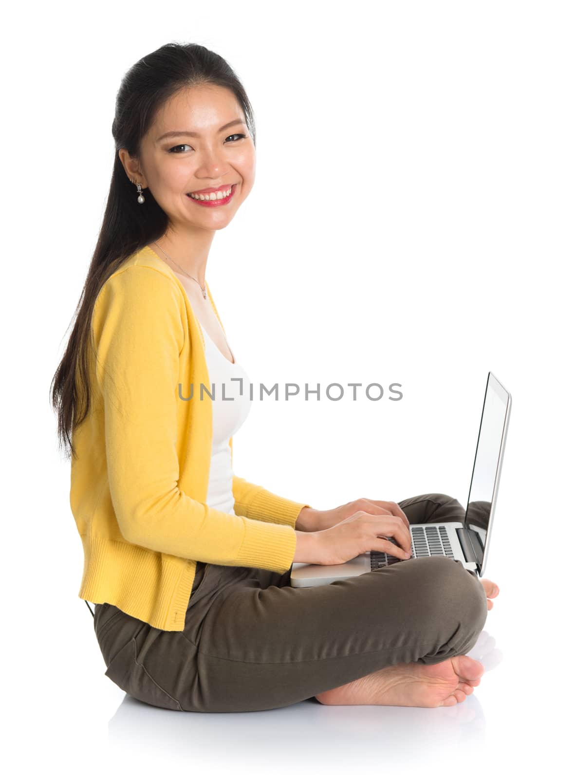 Side view full body Asian woman in yellow blouse using laptop computer notebook, looking at camera, seated on floor isolated on white background.