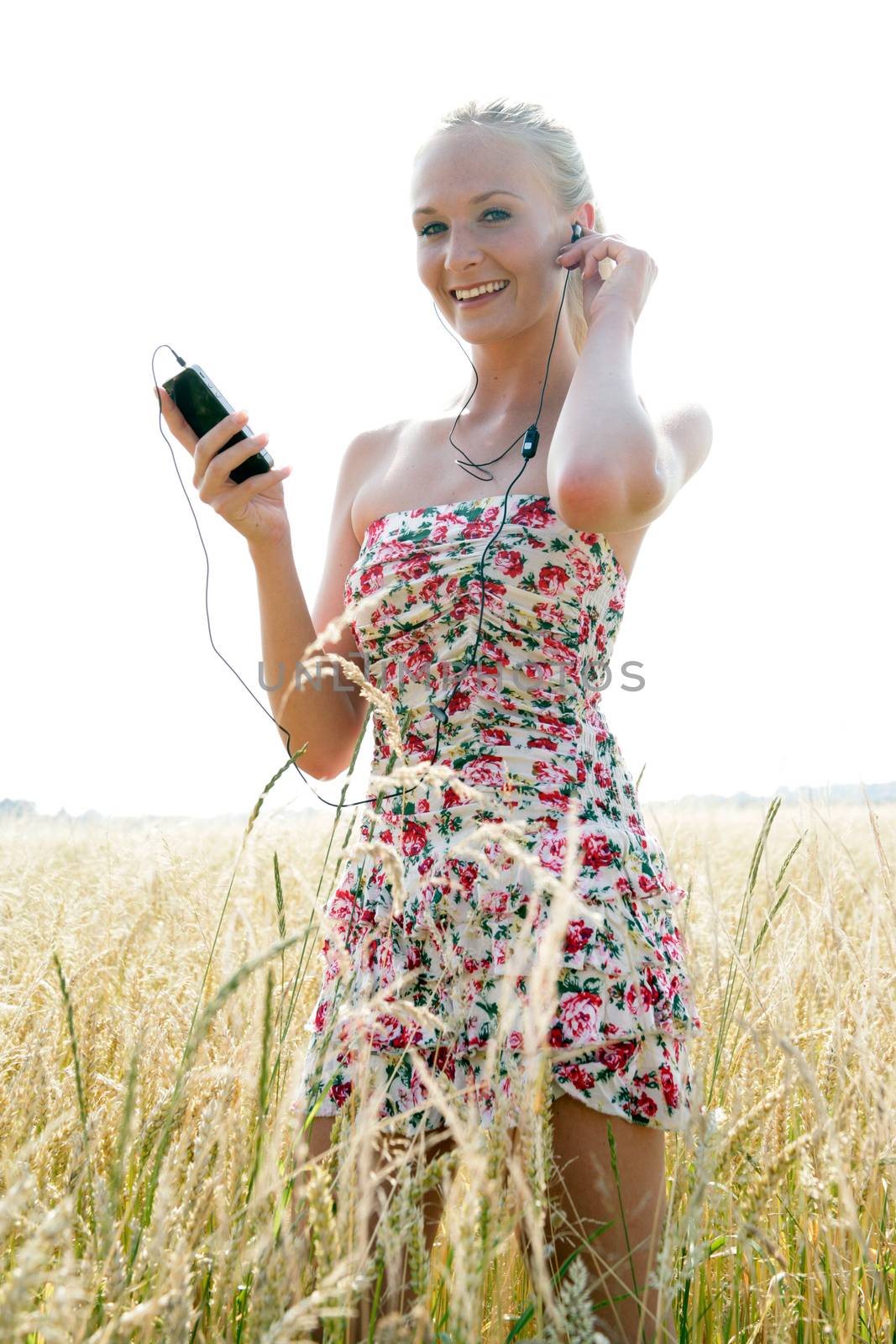 Young attractive woman is standing in a wheat field and listening to music with her smartphone. She looks happy and relaxed.