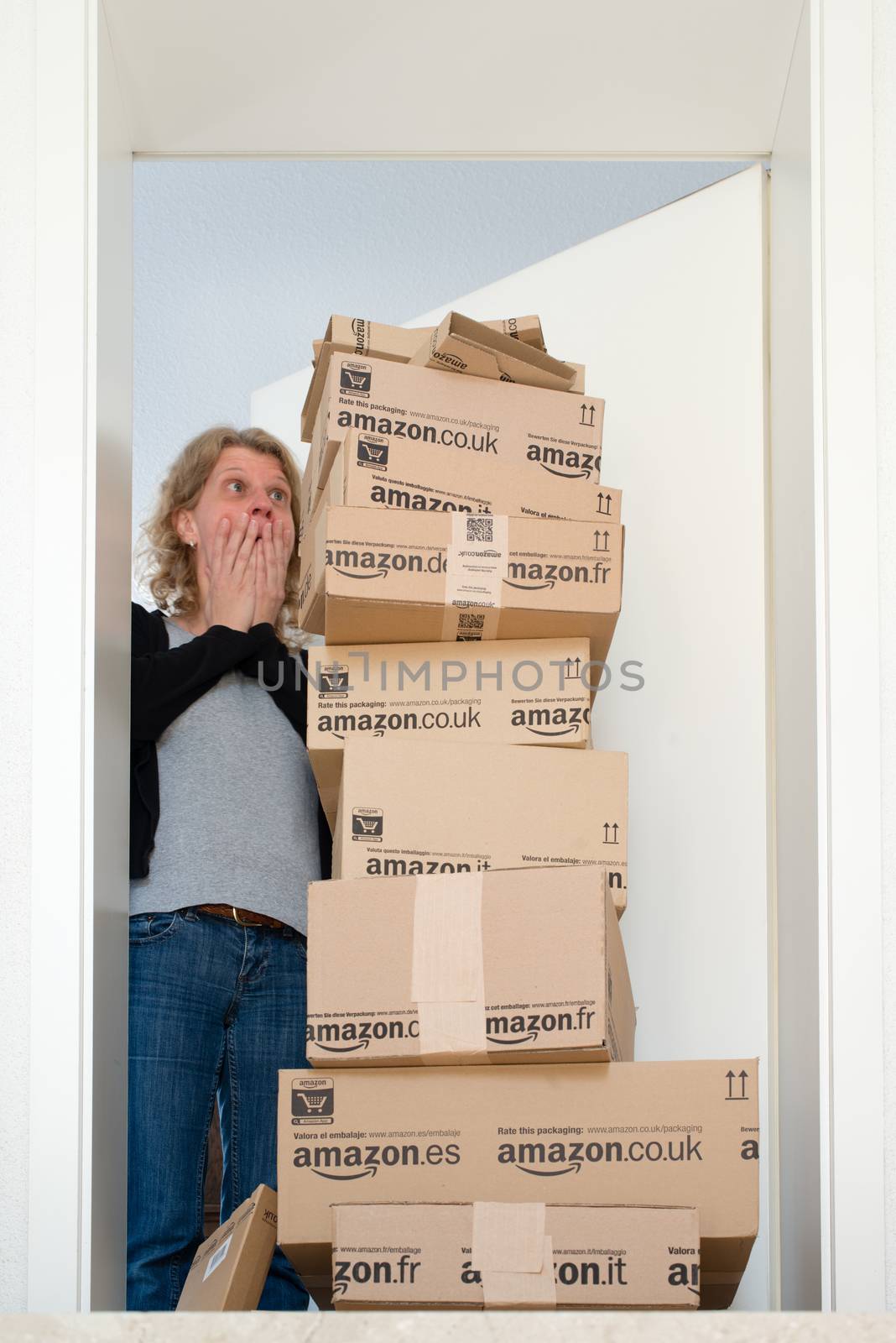 OSTFILDERN-SCHARNHAUSEN, GERMANY - MAY 18, 2014: A woman is horrified by a large stack of parcels by Amazon.com in different sizes waiting in front of the entrance door to her flat on May, 18, 2014 in Ostfildern-Scharnhausen near Stuttgart, Germany. This conceptual photo can serve different purposes: It might demonstrate the domination of Amazon.com in the area of online shopping or the trend in general to shop online for all the different items you need in your personal life.