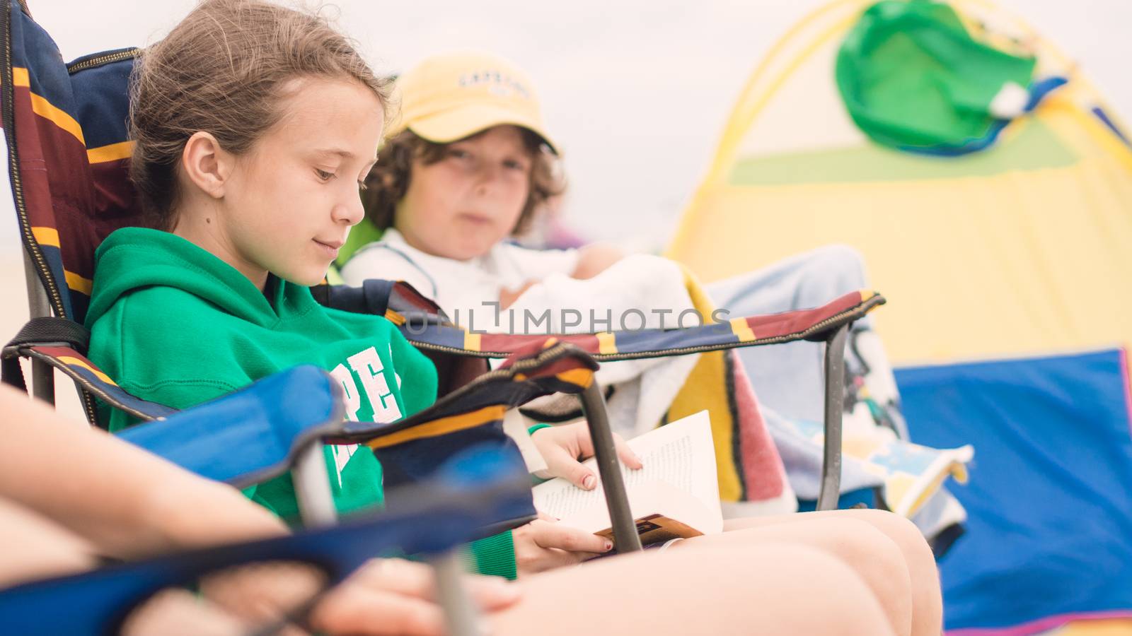 Young girl reading in a chair on the beach while is brother is watching