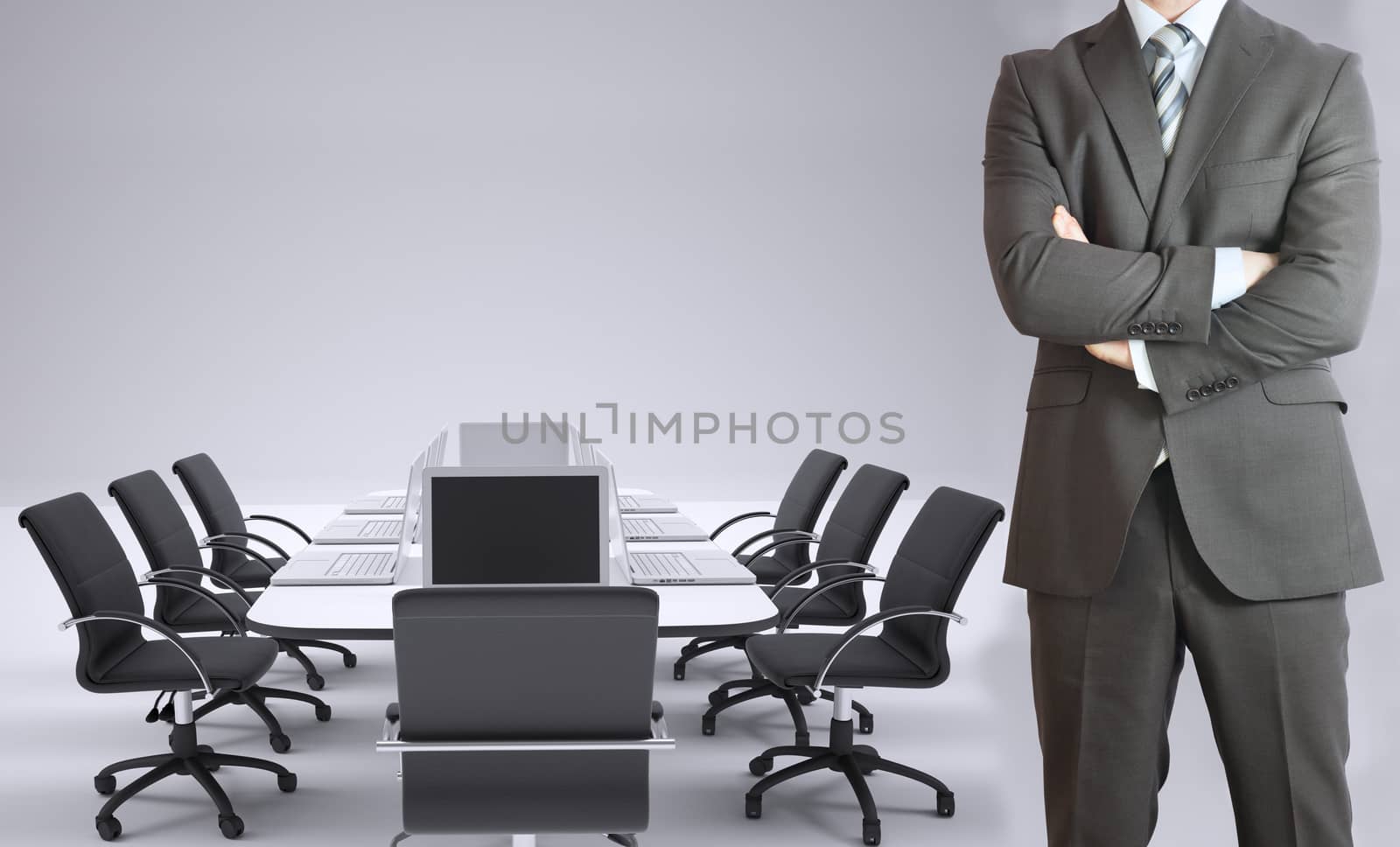 Businessman standing with his arms crossed. Conference table, chairs and laptops as backdrop
