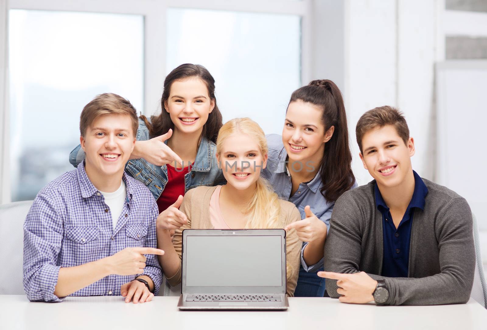 education, technology, advertisement and internet concept - group of smiling students pointing to blank black laptop screen