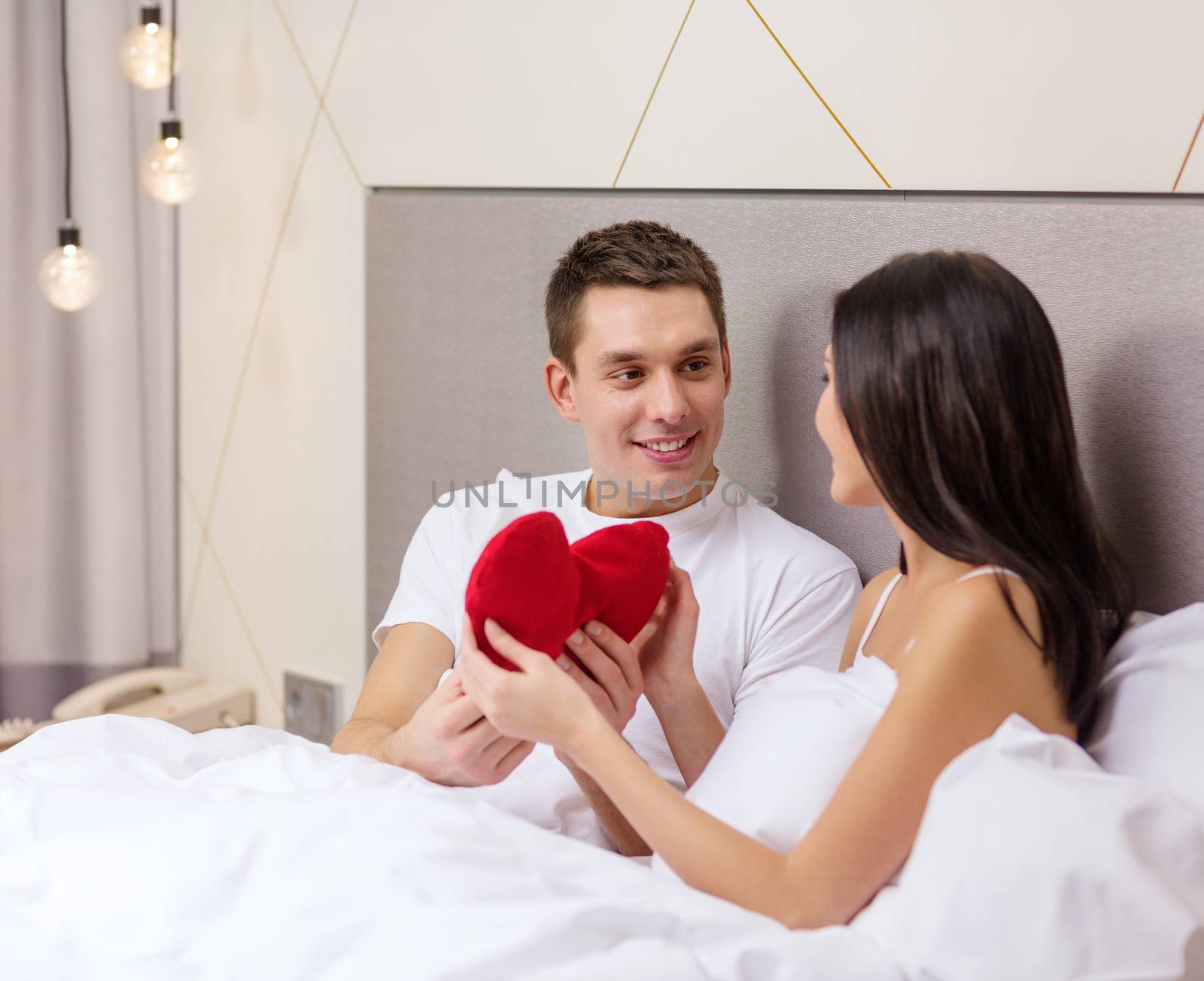 hotel, travel, relationships, holidays and happiness concept - smiling couple in bed with red heart-shaped pillow