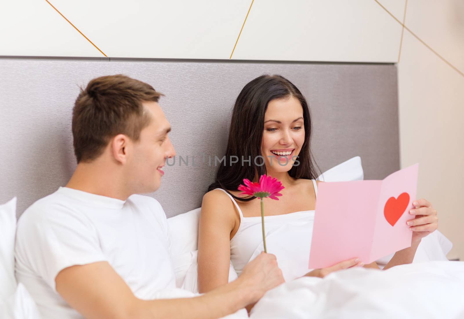 hotel, travel, relationships, holidays and happiness concept - smiling couple in bed with postcard and pink flower