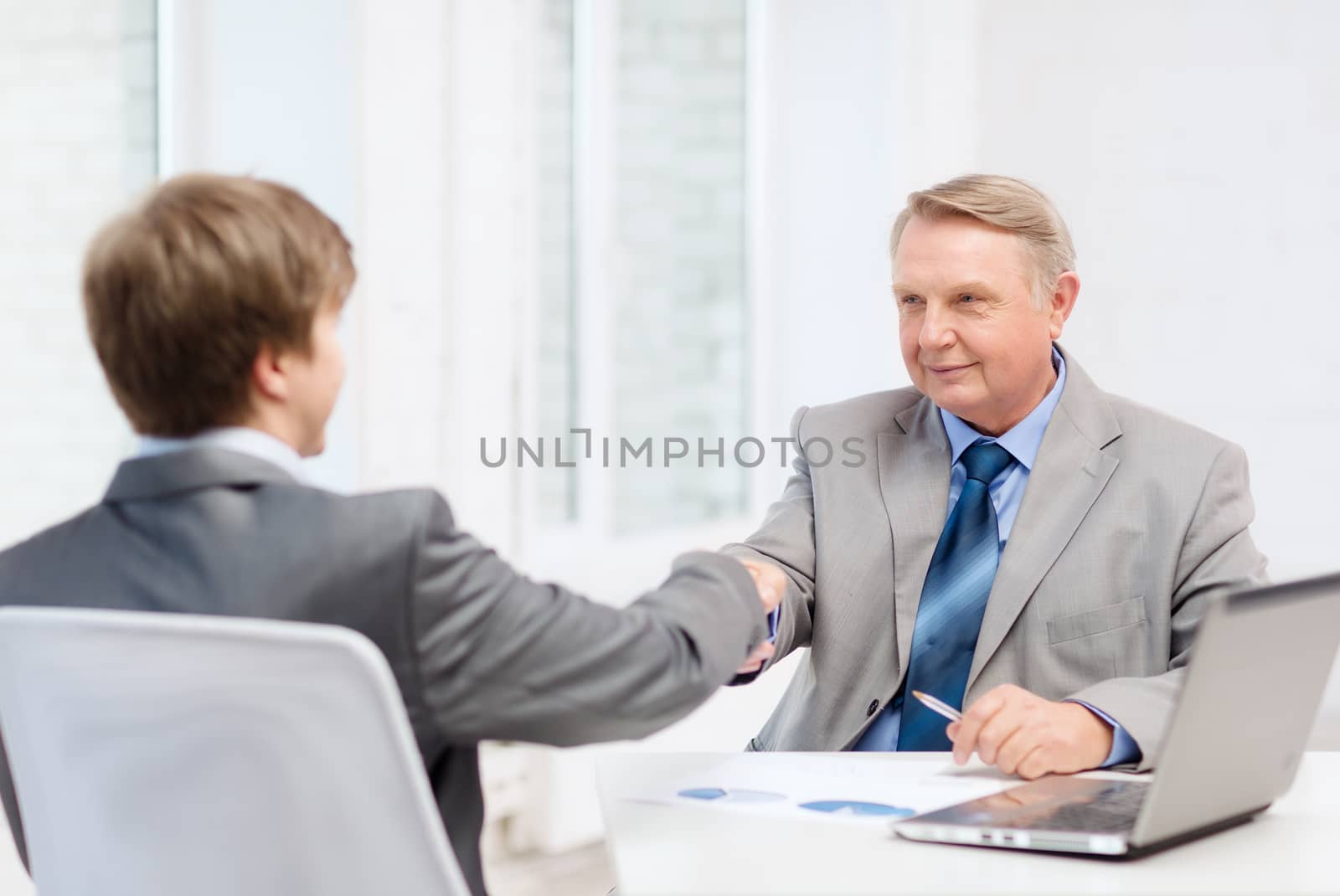 business, technology and office concept - older man and young man shaking hands in office