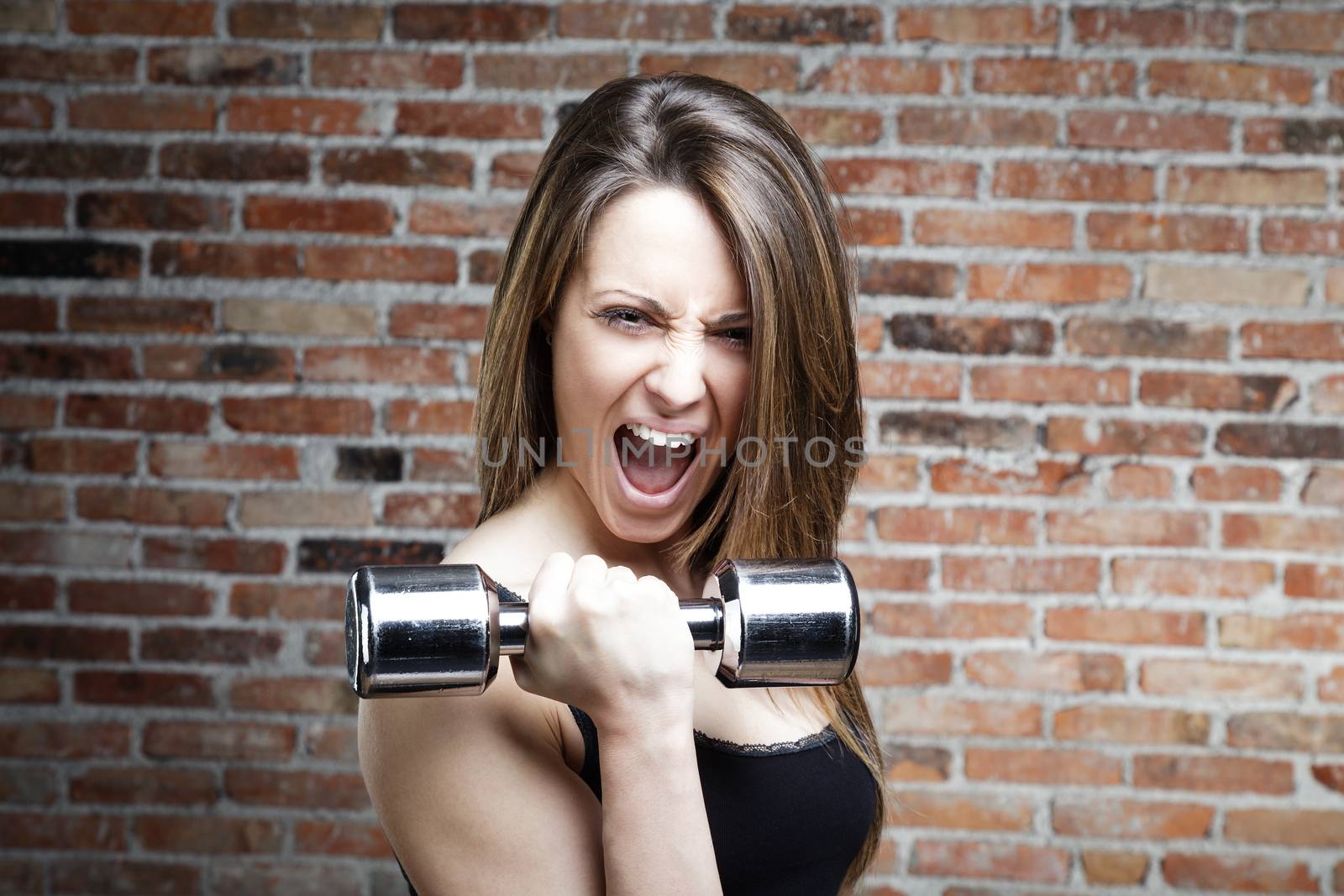 Portrait of Young shouting fit woman lifting dumbbells on brick background