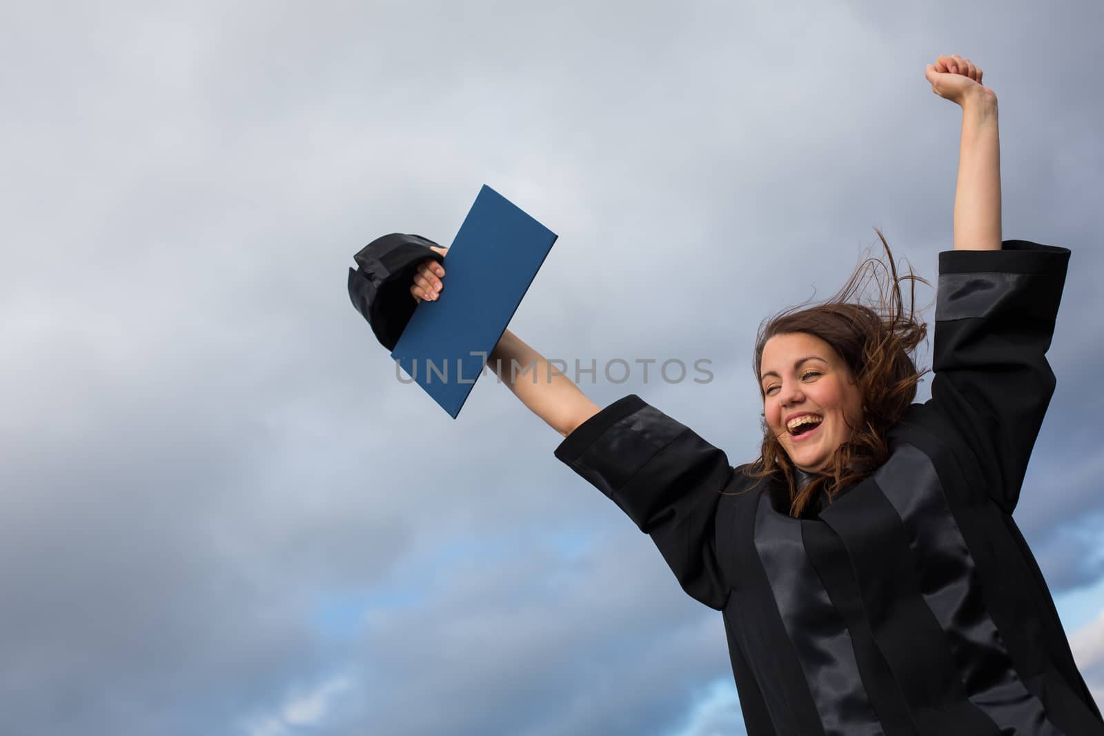 Pretty, young woman celebrating joyfully her graduation - spreading wide her arms, holding her diploma, savouring her success (color toned image; shallow DOF)