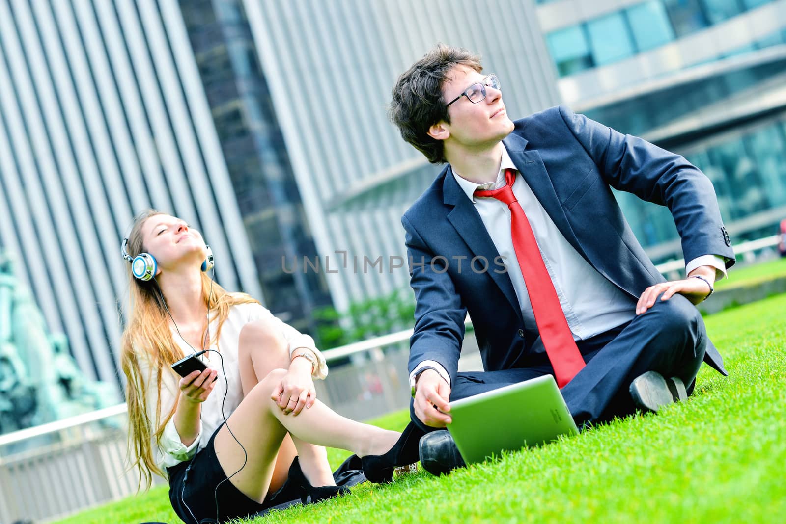 Successful Junior executives dynamics laying down on green grass near a modern glass office building, listening music and using laptop