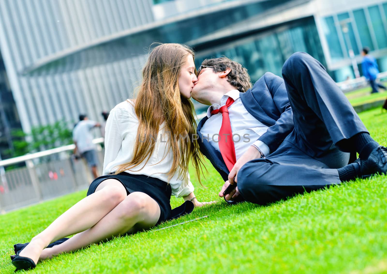 Successful lovers laying down on green grass near a modern glass office building, listening music and using laptop