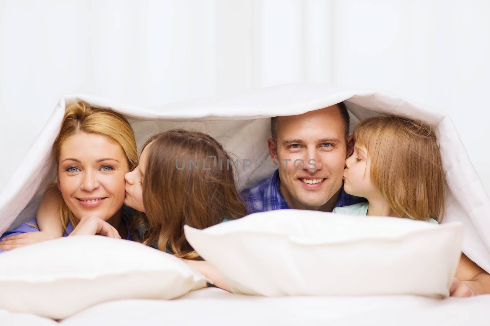 family, children and home concept - two little girls kissing their parents on cheek under blanket at home