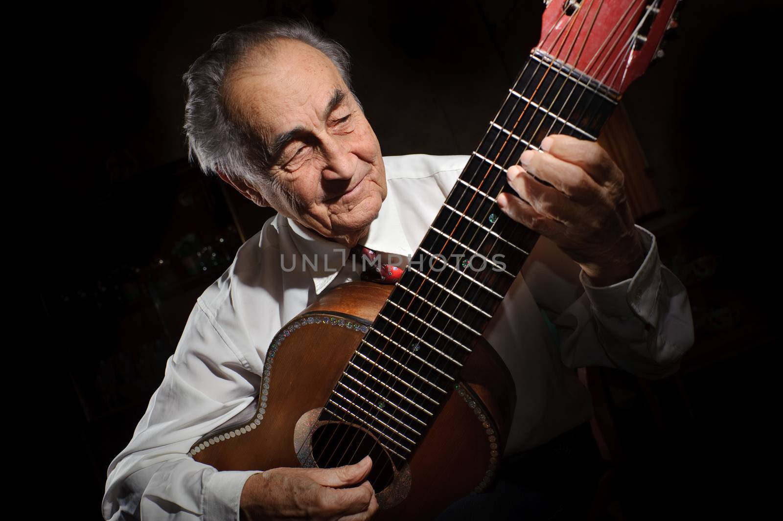 An elderly man in white shirt playing an acoustic guitar. Dark background. 