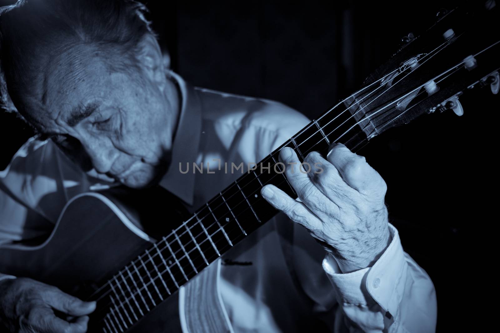 An elderly man in white shirt playing an acoustic guitar. Dark background. Monochrome.  Focus on the hand.