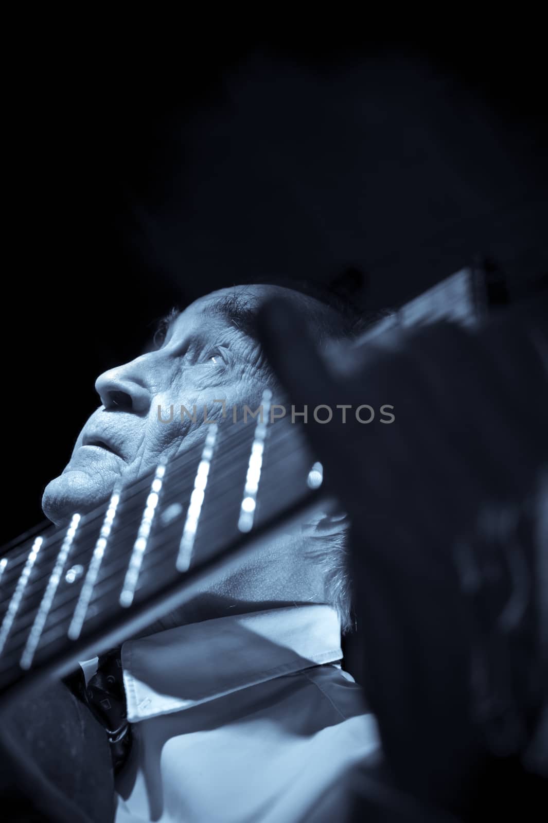 An elderly man in white shirt playing an acoustic guitar. Dark background. Monochrome.